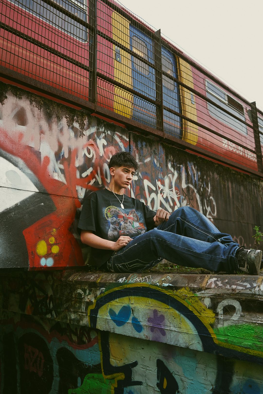 Young man in graphic tee and jeans sitting on a ledge against a colorful graffiti-covered wall outdoors.