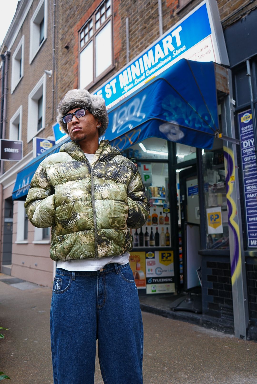 A man in a camo puffer jacket and fur hat stands outside a minimart on a city street.
