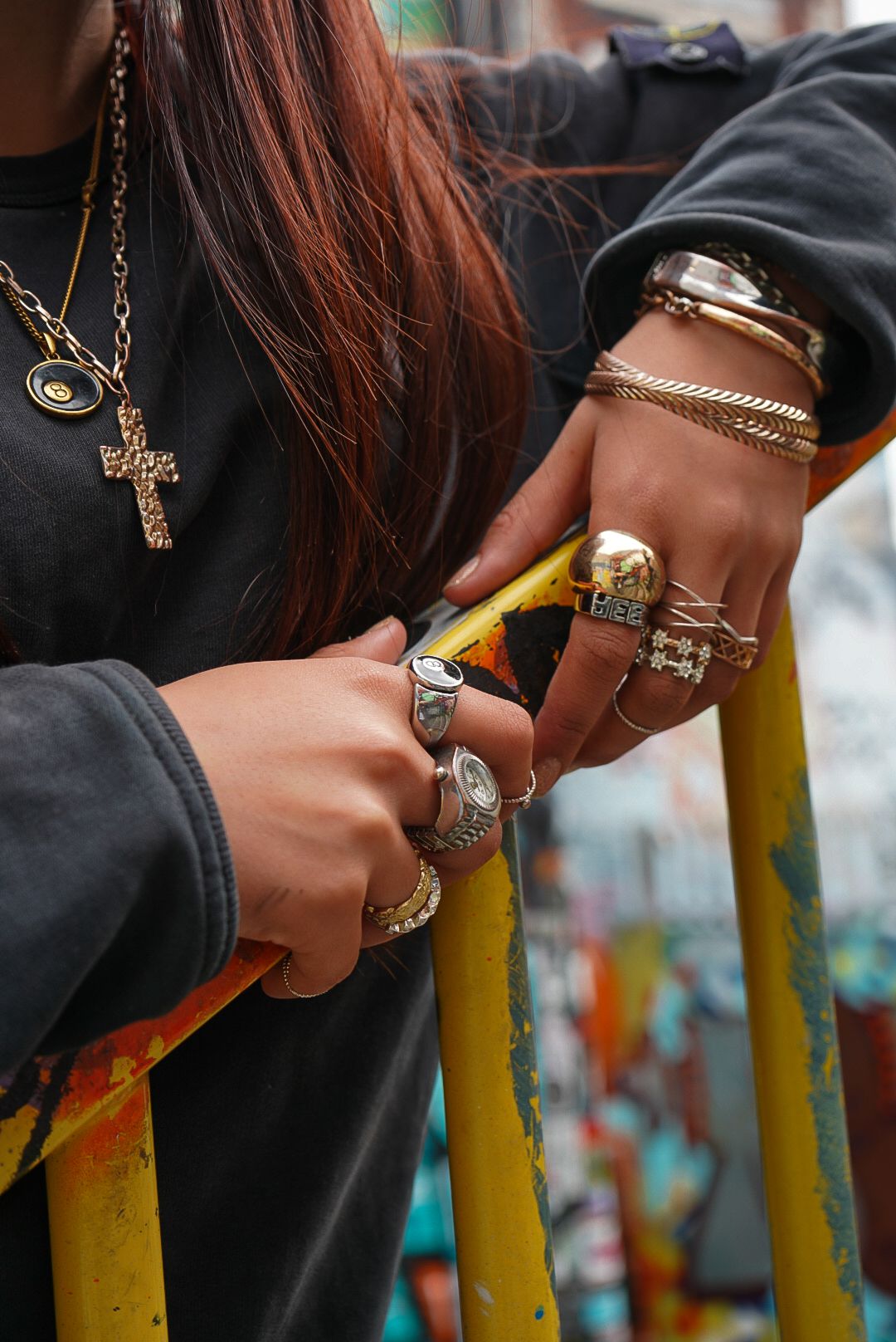 Close-up of two people's hands adorned with gold and silver rings, bracelets, and cross necklace on yellow railing.