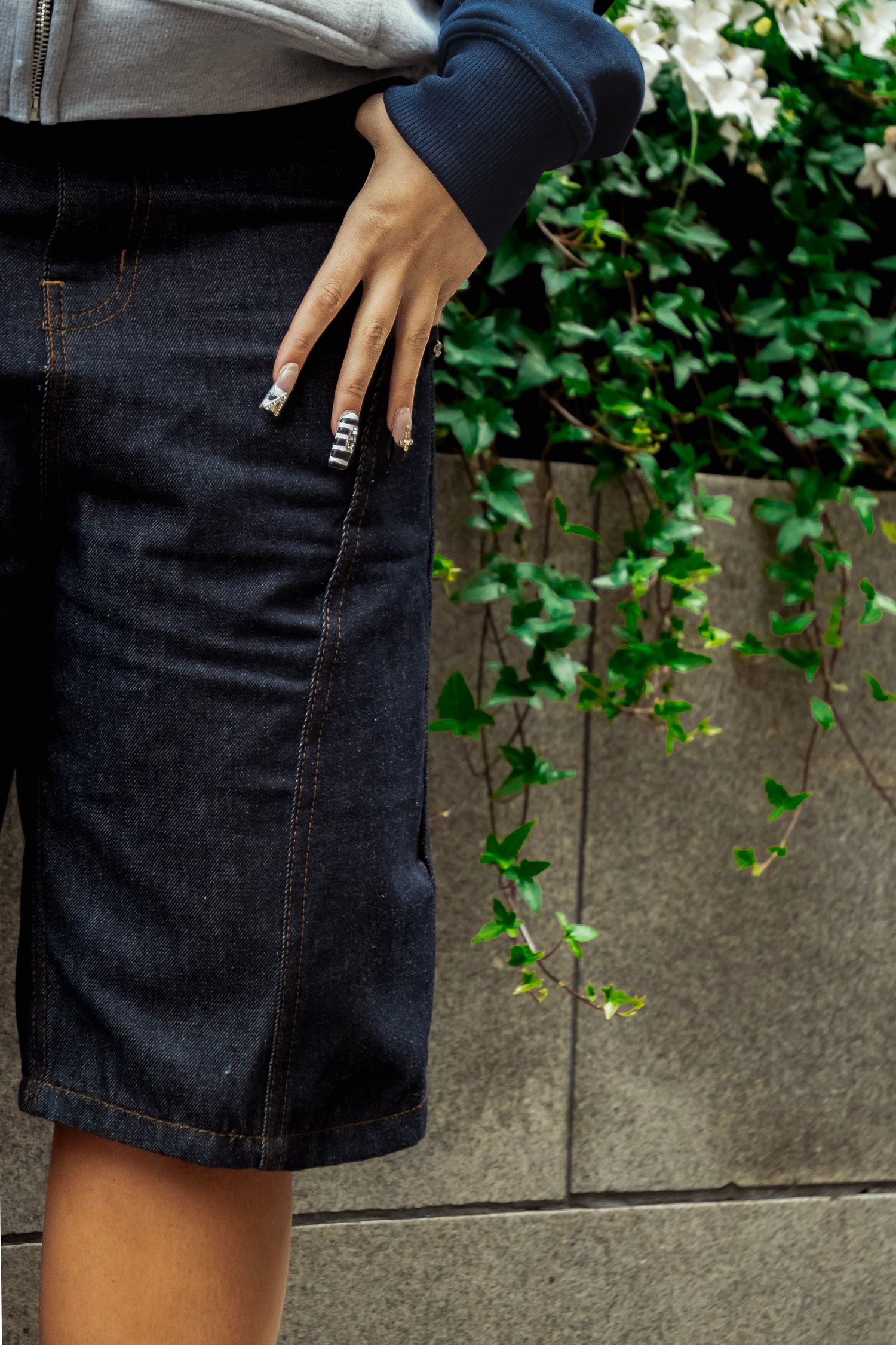 Woman wearing dark denim knee-length shorts with hand in pocket, standing near a stone wall with ivy.