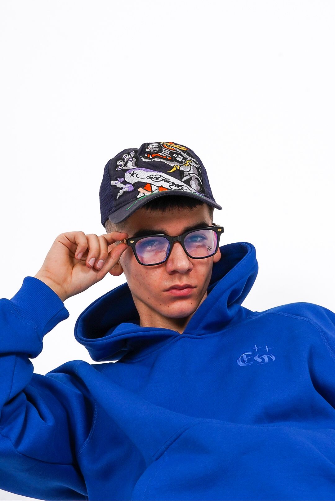 Young man in royal blue hoodie adjusting graphic cap while wearing clear-framed glasses.