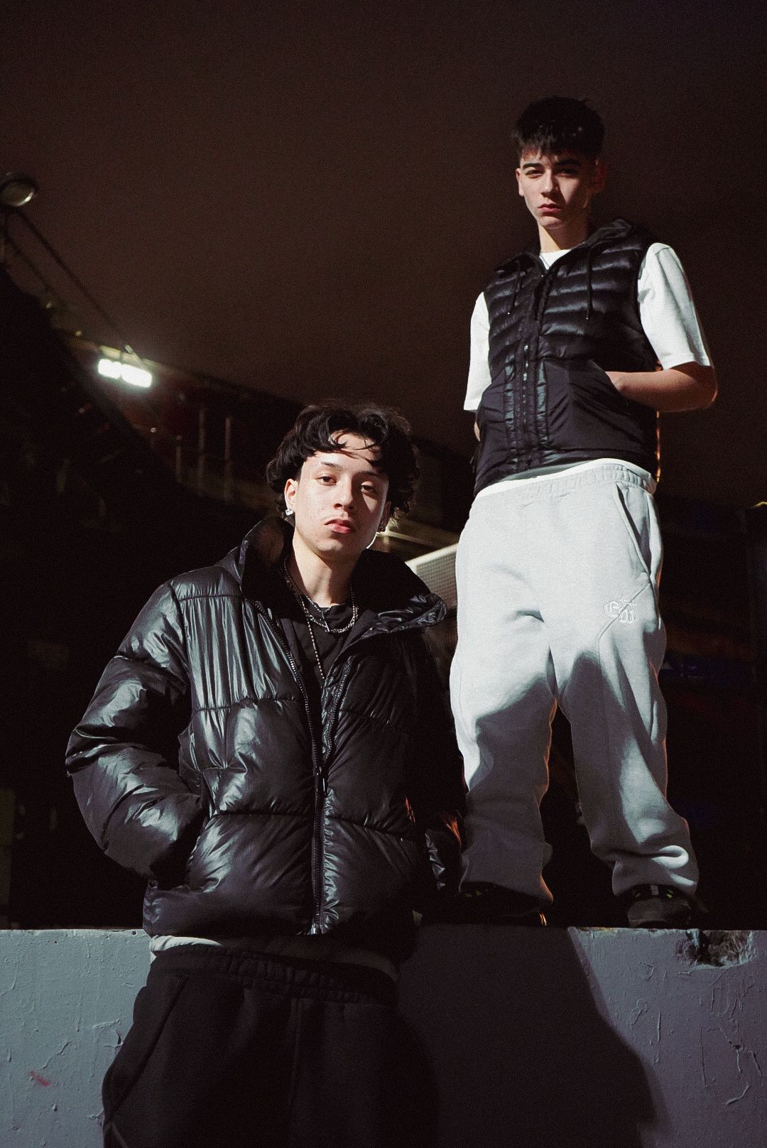 Two young men in black puffer jacket and white vest posing outdoors at night under floodlights.