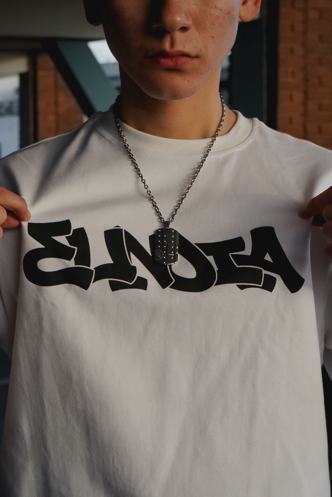 Young man wearing a white t-shirt with ELNDIA graffiti logo and silver chain necklace.
