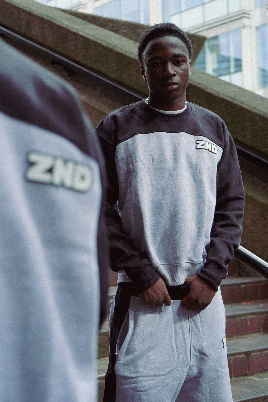 Young man wearing ZND branded grey and dark colorblock crewneck sweatshirt on urban steps.