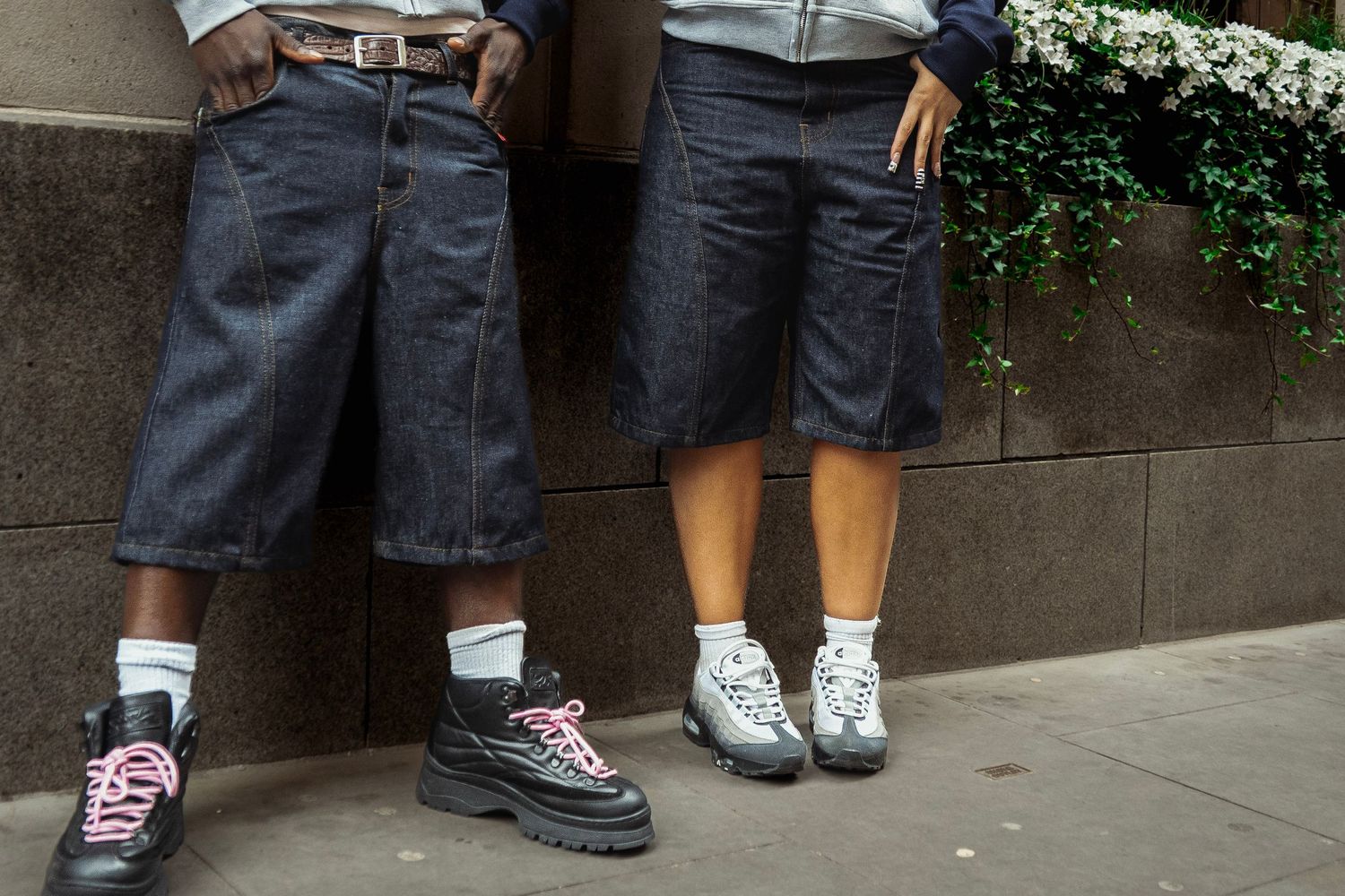 Two people wearing dark denim shorts and chunky sneakers leaning against a concrete wall outdoors.