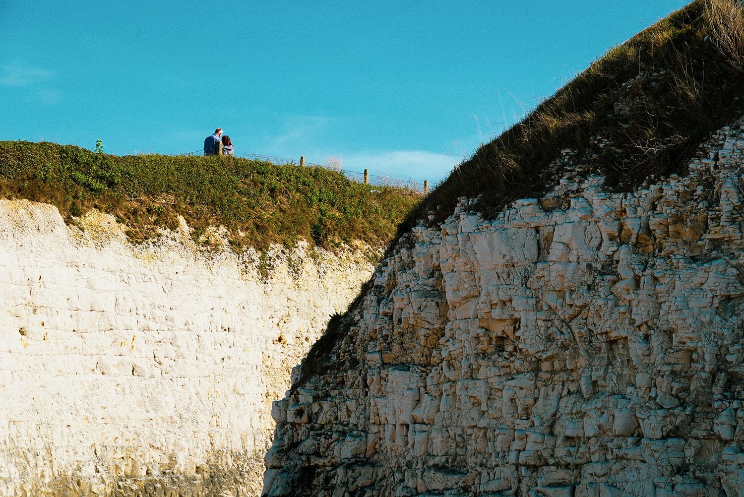 Two people stand atop white chalk cliffs covered in green vegetation against a bright blue sky.