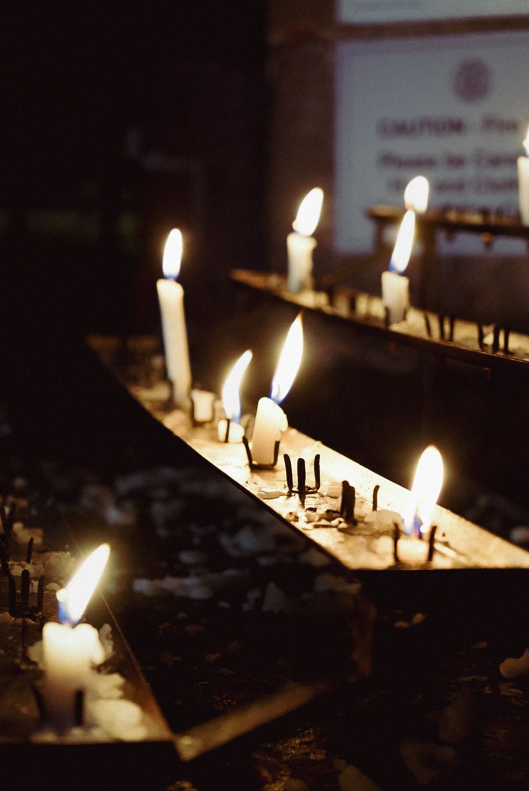 Rows of white candles burning on a wooden rack in a dark church or memorial setting.