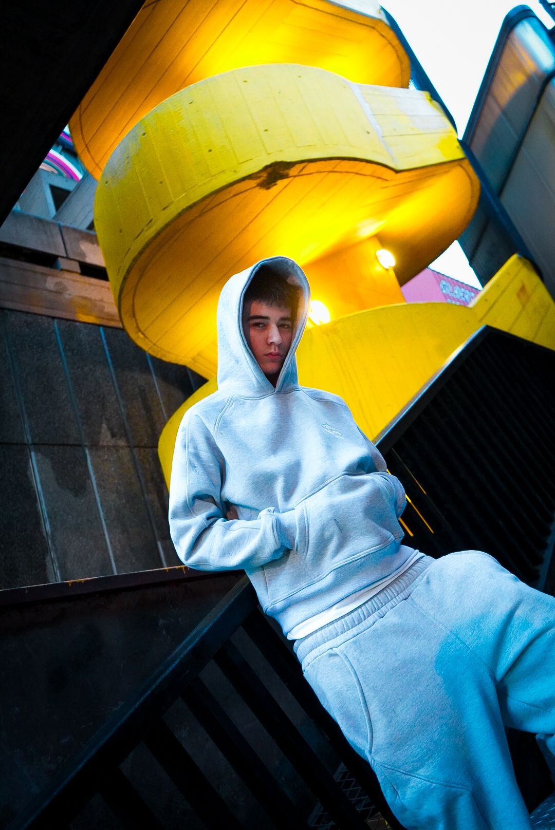 Young man in white hoodie and sweatpants poses on dark stairs with vibrant yellow spiral structure behind him.