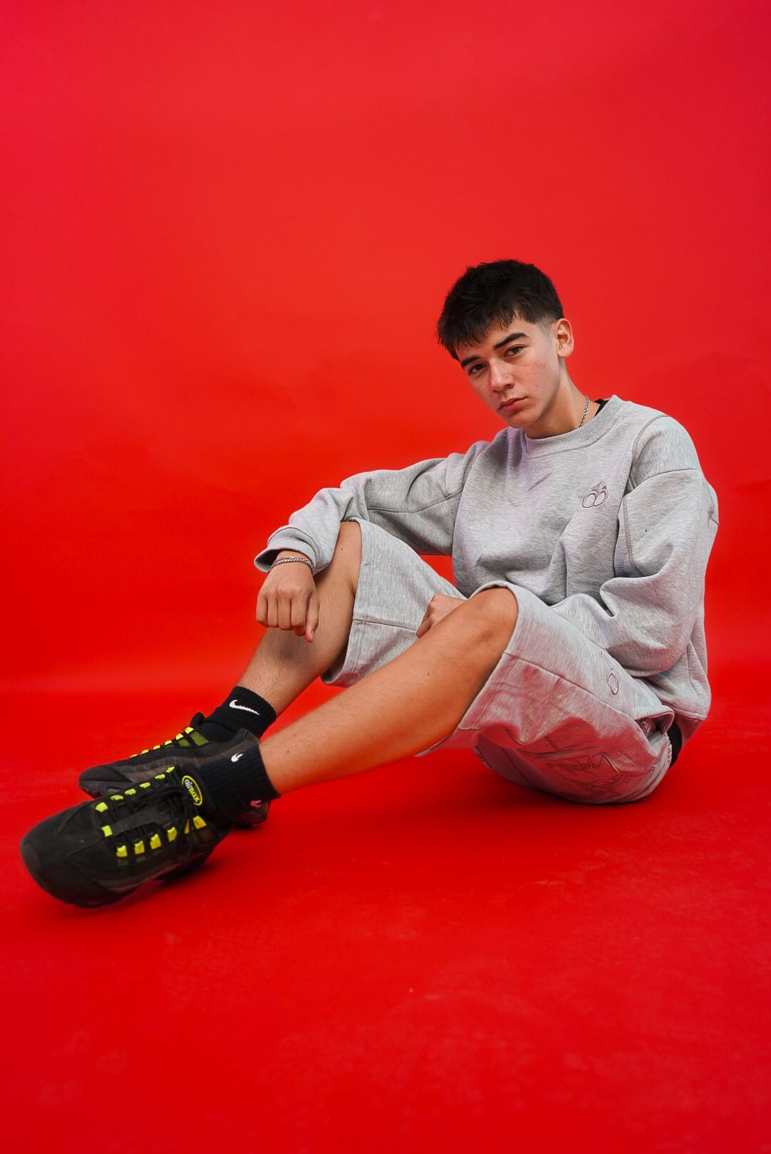 Young man in grey oversized sweatsuit sits on floor against a bold red background wearing black sneakers.