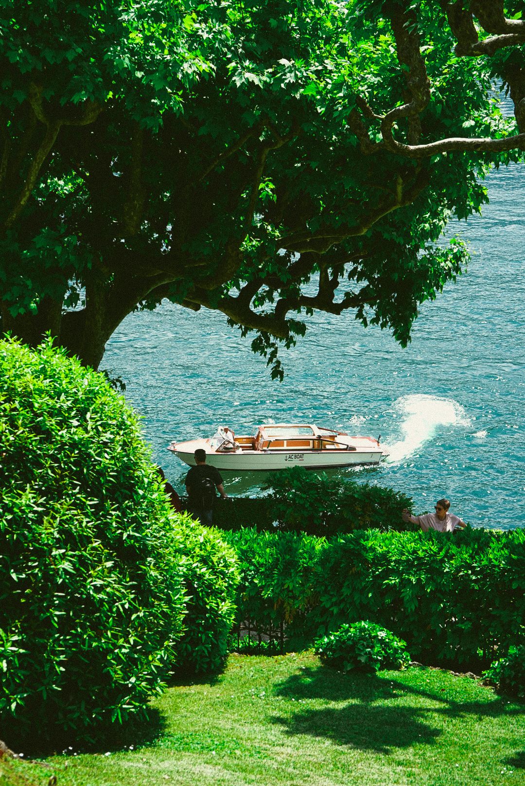 White vintage speedboat cruising on blue lake waters framed by lush green trees and manicured garden.