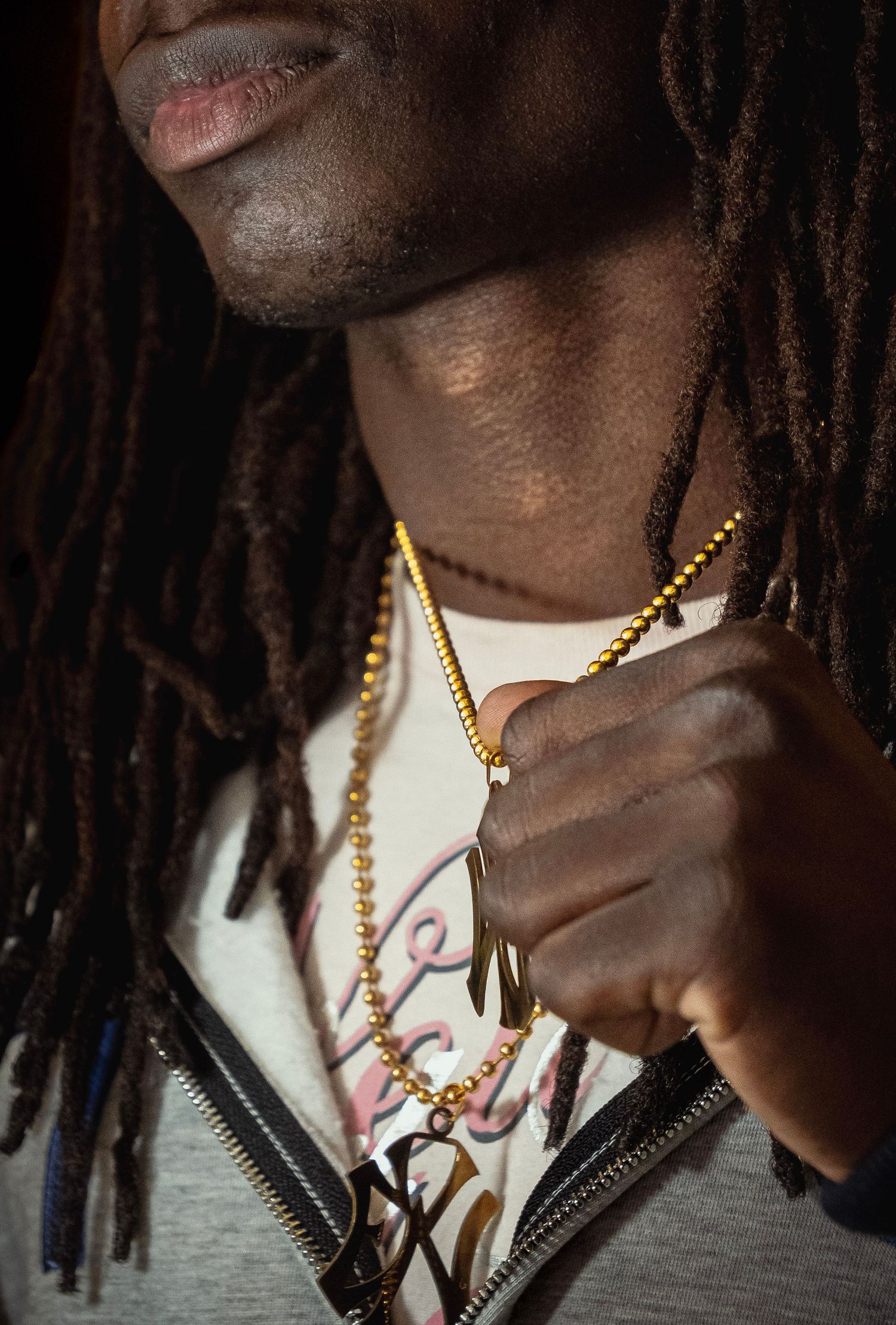Black man with dreadlocks wearing gold chain necklaces and a white graphic tee with a zip-up jacket.