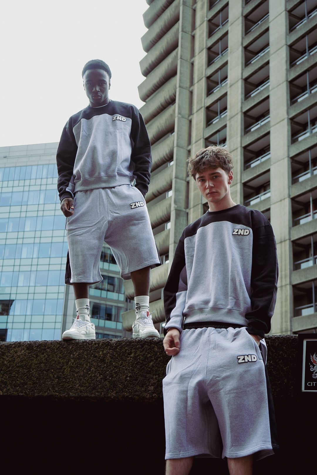Two young men wearing ZND grey and black colorblock sweatshirts and shorts outside urban tower block.