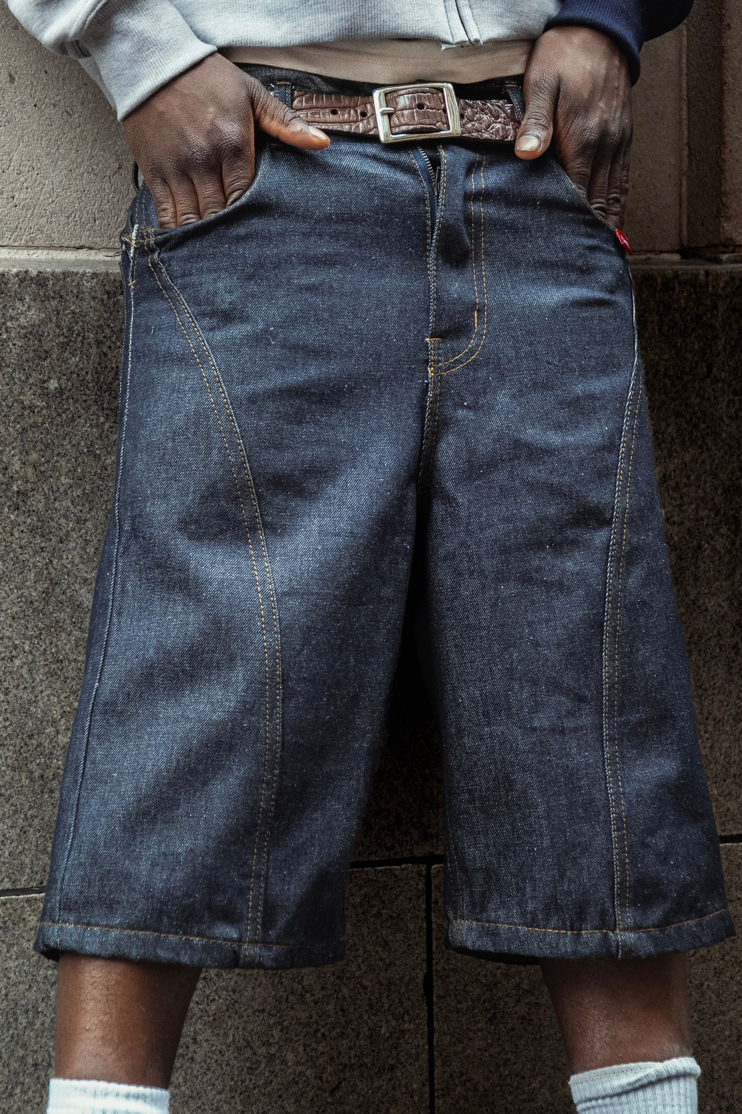 Person wearing oversized dark denim shorts with brown belt and white socks, standing against a concrete wall.