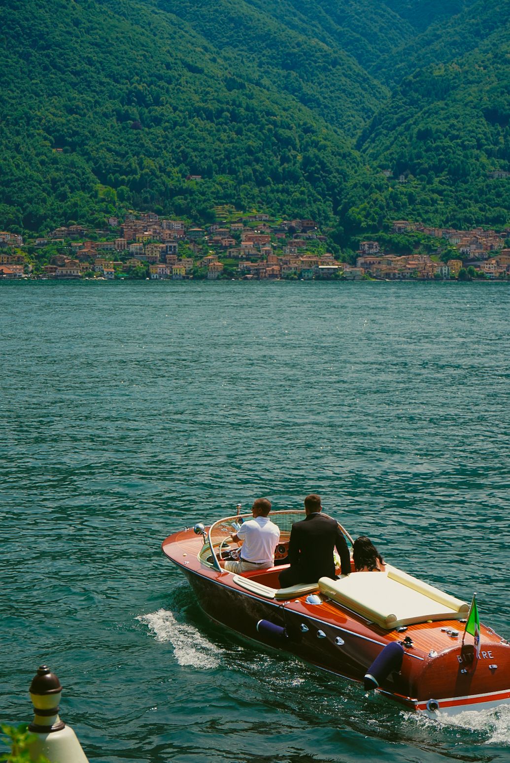 Three people riding classic red wooden motorboat across turquoise alpine lake with hillside village beyond.