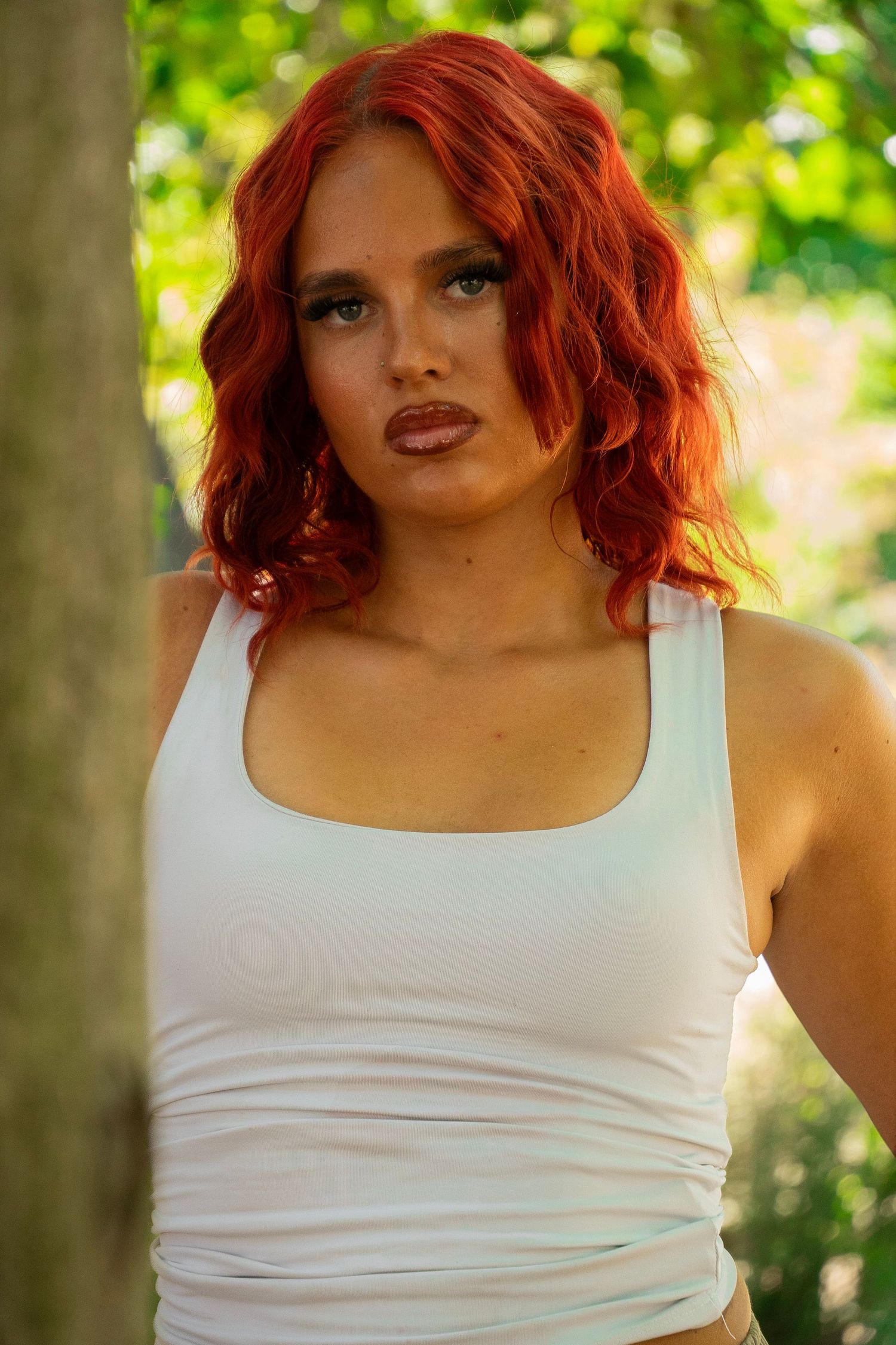 Young woman with vibrant red wavy hair wearing a white tank top, posing outdoors near trees.