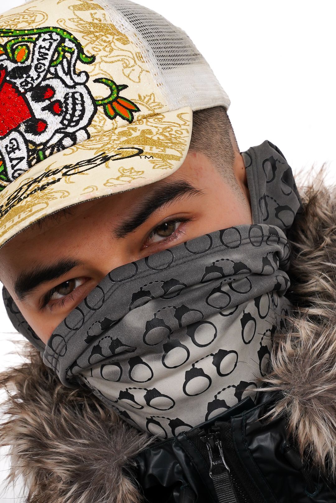 Young man wearing decorative Ed Hardy cap and grey skull-print face covering with fur-lined jacket close-up portrait.