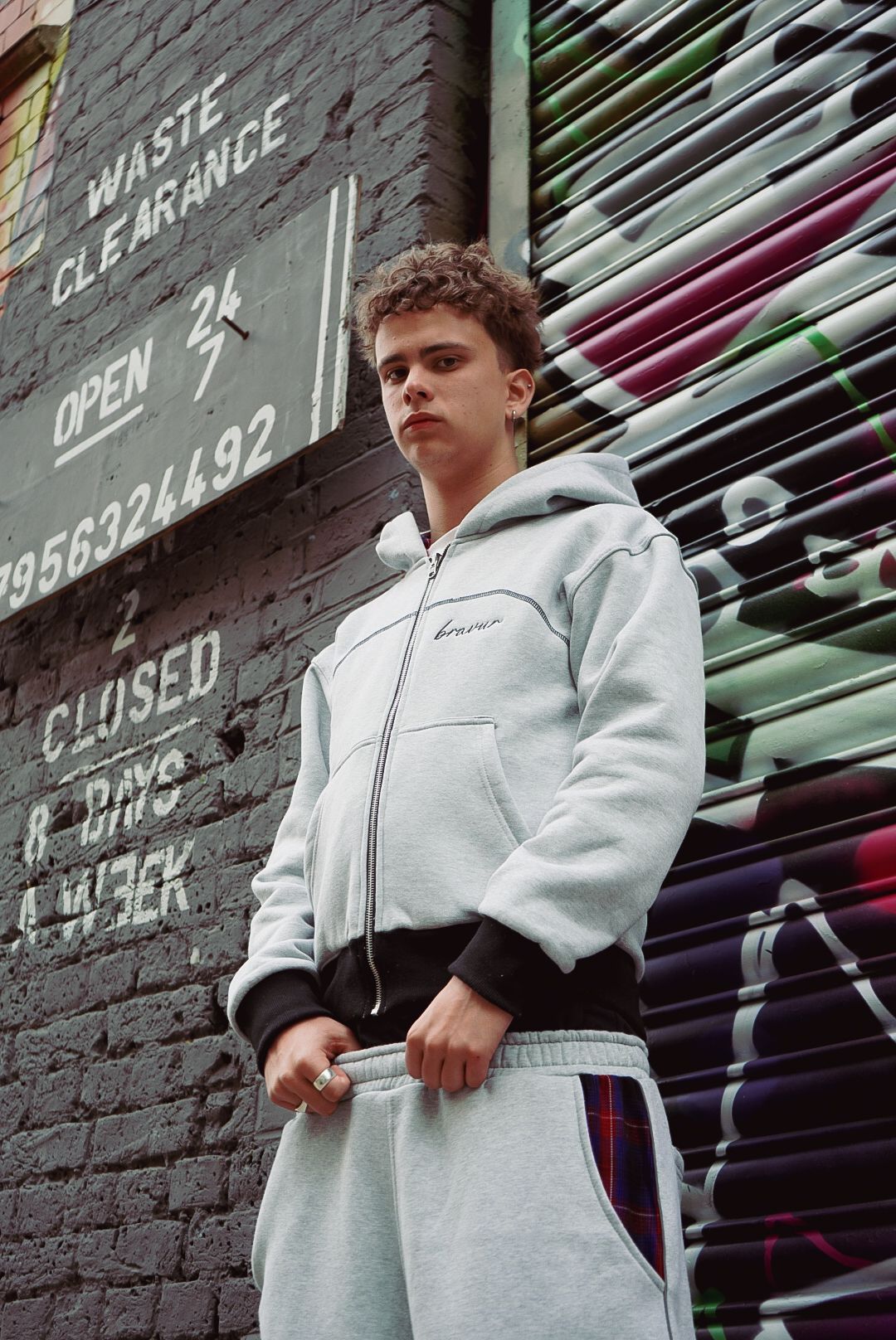 Young man in grey zip-up hoodie and sweatpants stands by graffiti wall next to a waste clearance sign.