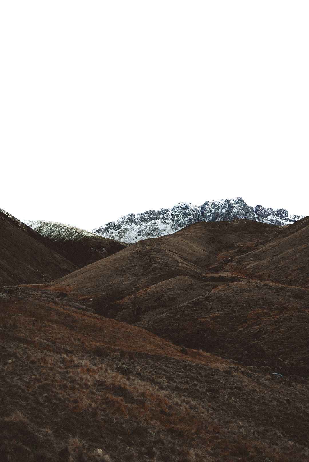 Dramatic landscape of brown rolling hills with snow-capped mountain peaks against white sky.