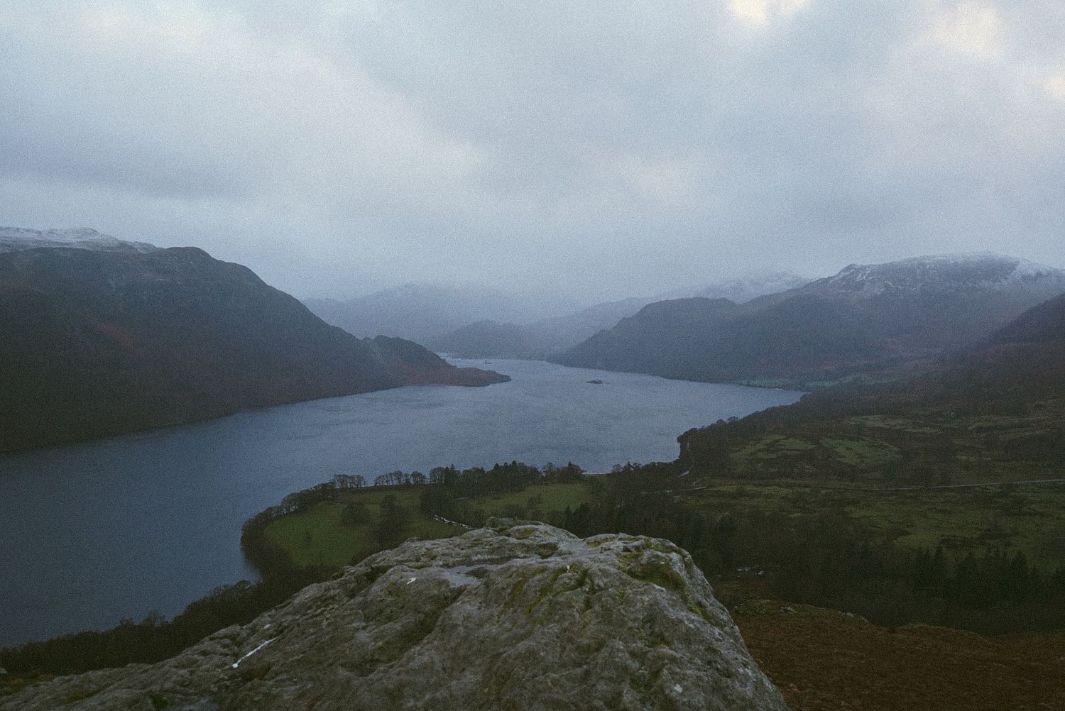 Aerial view of a long lake between misty snow-dusted mountains under cloudy sky.