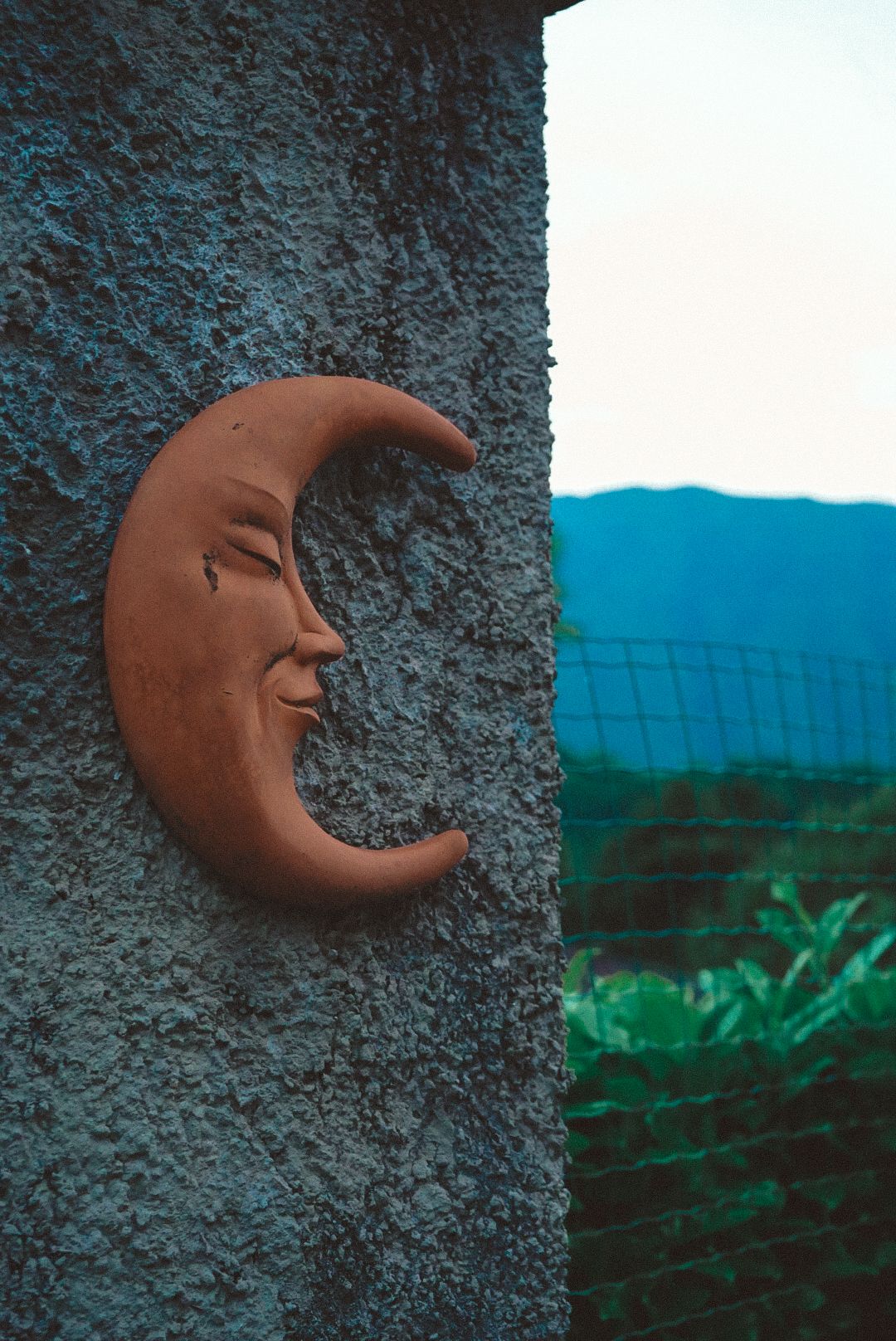 Terracotta crescent moon face sculpture mounted on textured grey wall with mountain landscape in background.