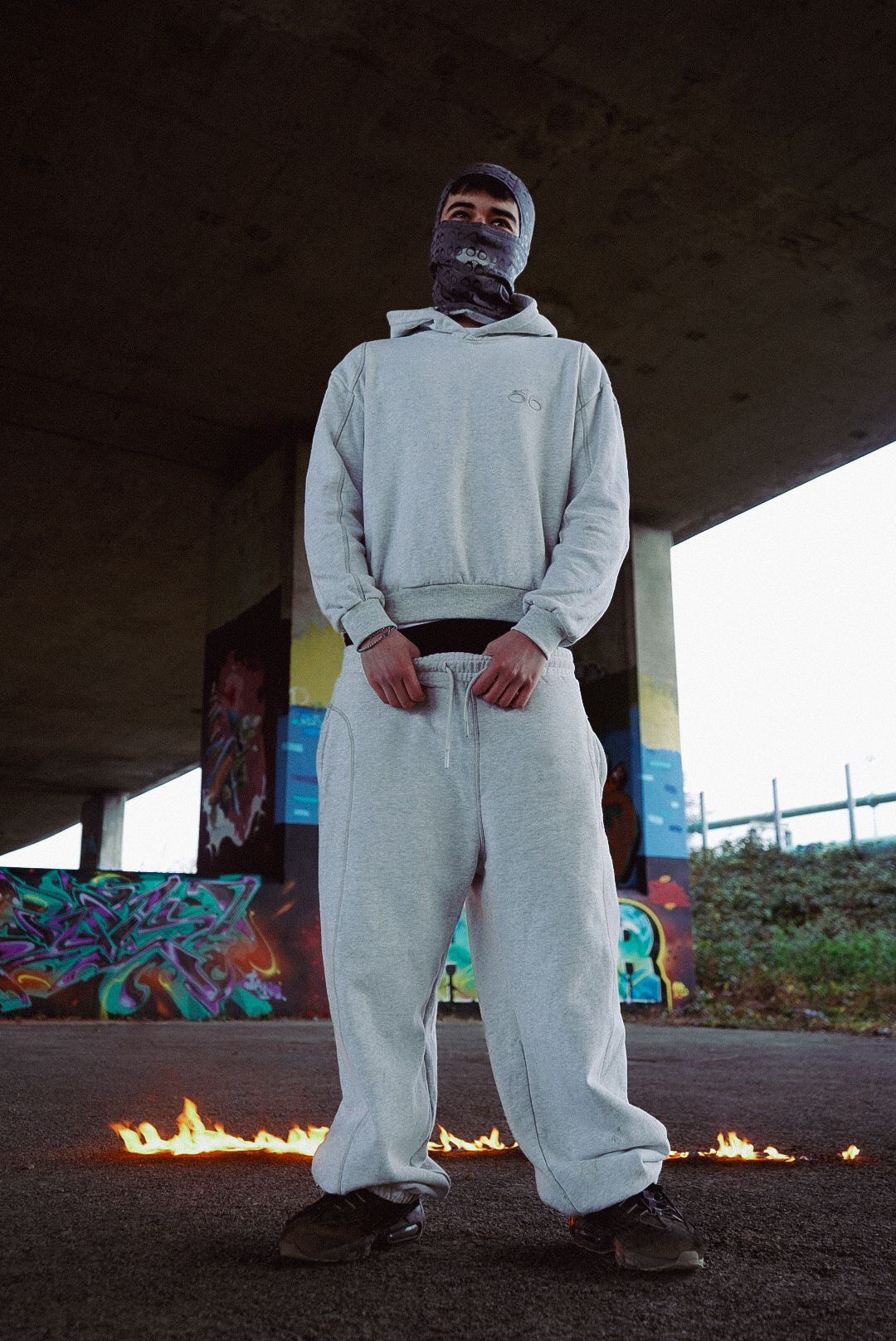 Person in grey hoodie and sweatpants standing before graffiti wall with flames on ground.