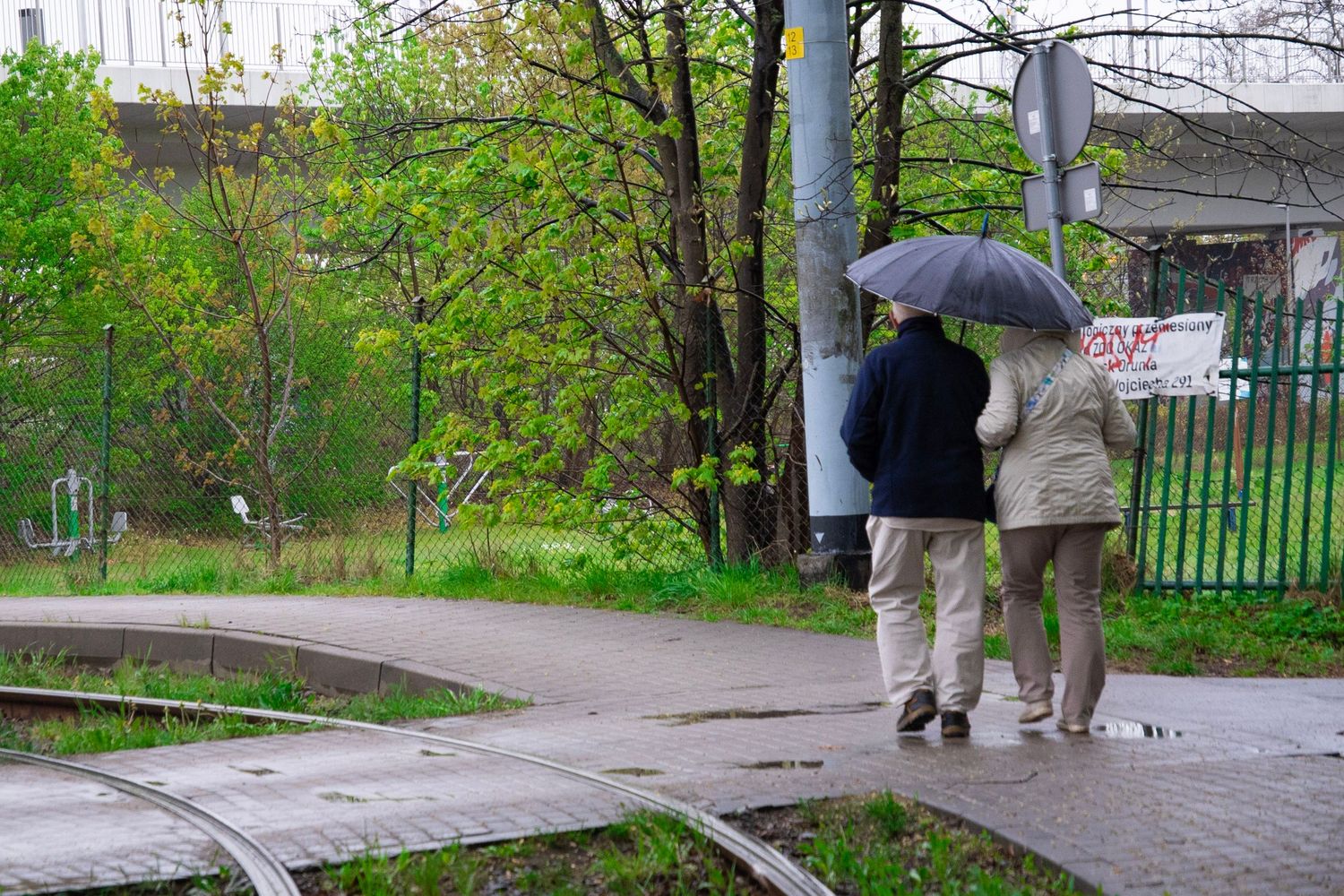 Two people sharing an umbrella walk along a rainy urban path surrounded by green trees and a metal fence.