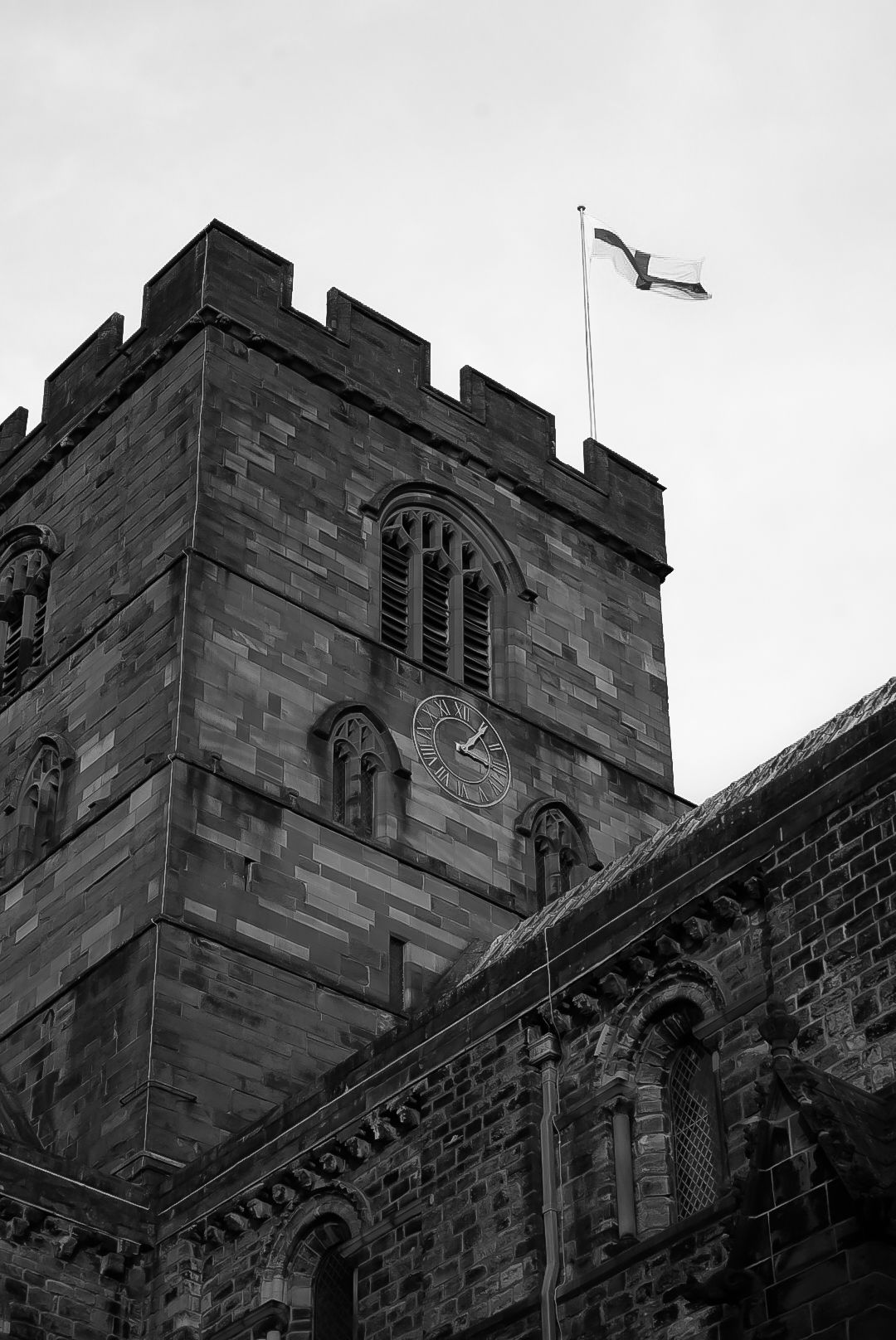 Black and white photo of a medieval stone church tower with a flag flying against grey sky.