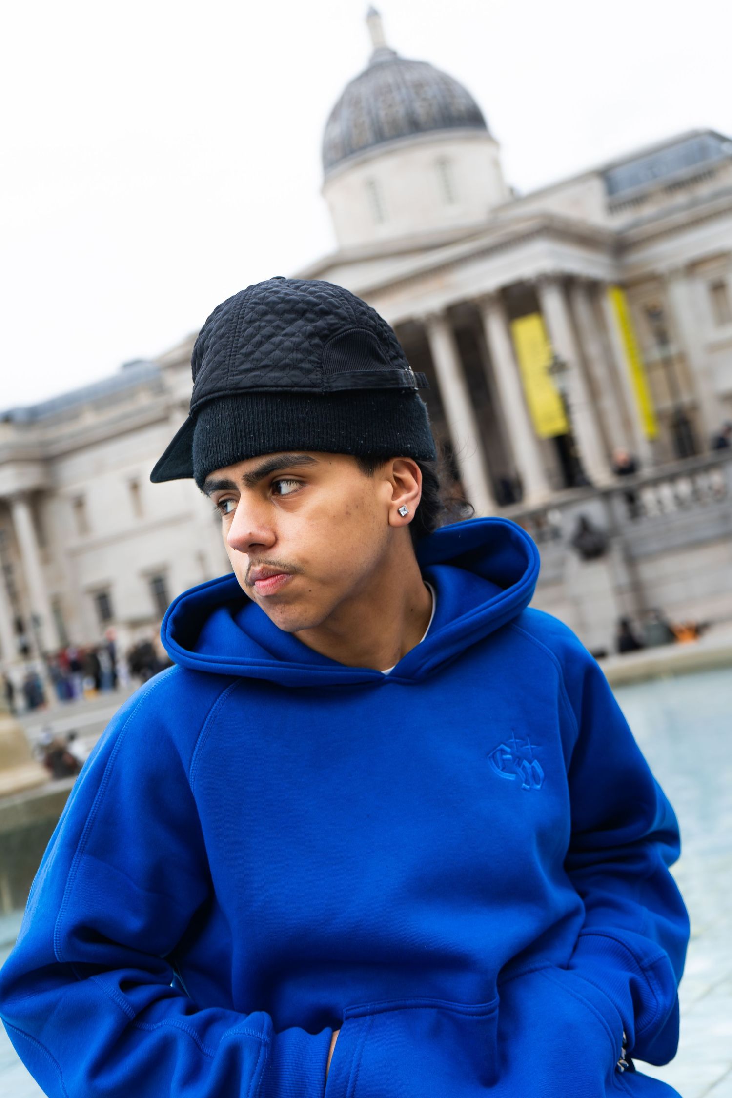Young person in blue hoodie and black cap posing outside the National Gallery in Trafalgar Square.