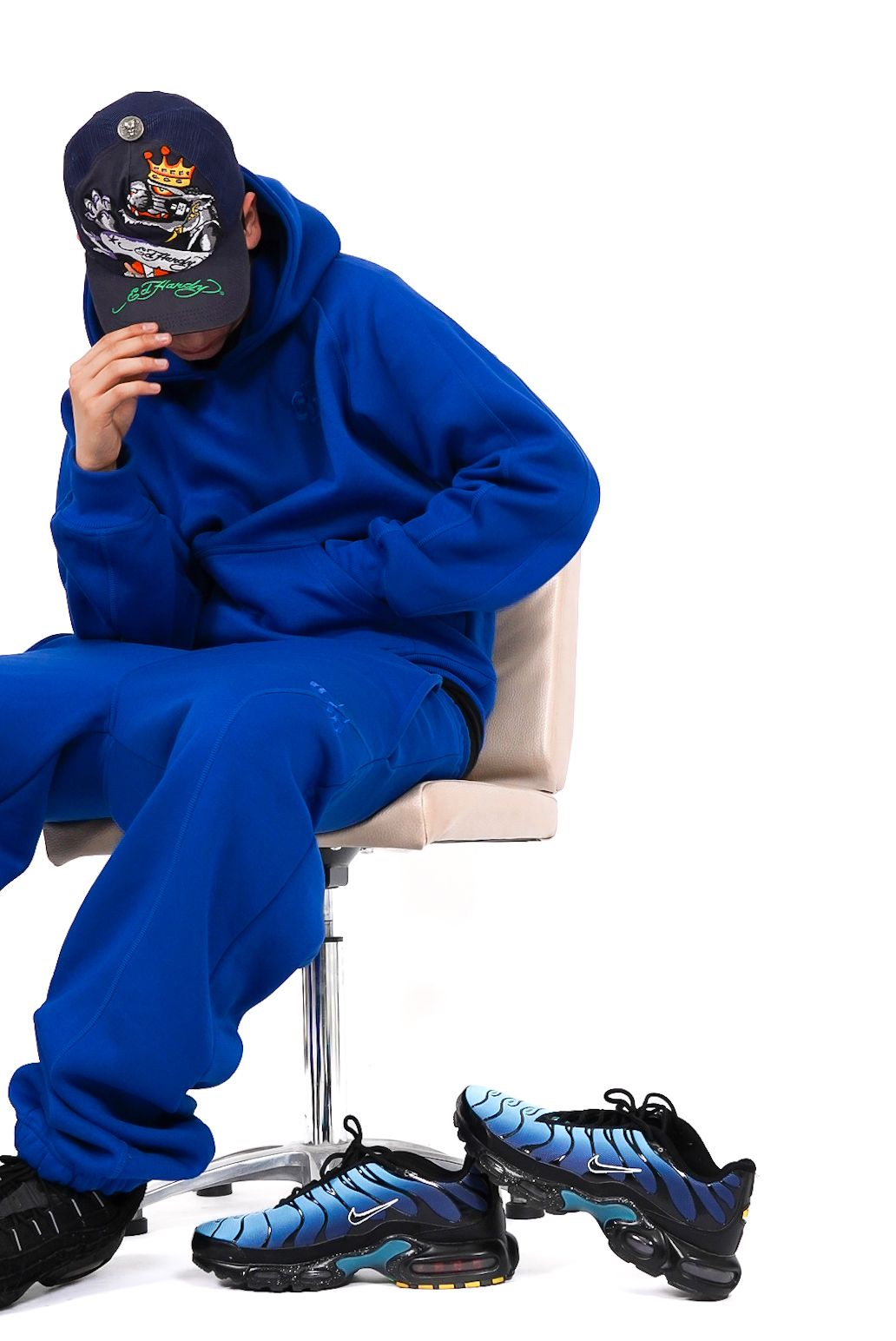 Person in blue hoodie and sweatpants sitting on chair, hiding face under navy cap, with Nike Air Max Plus sneakers.
