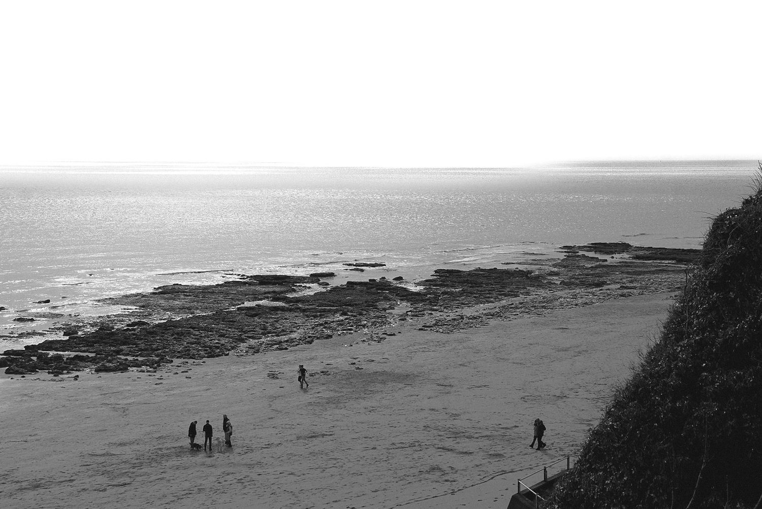 Black and white coastal beach scene with small figures walking on sandy shore beside rocky outcrops and calm ocean.