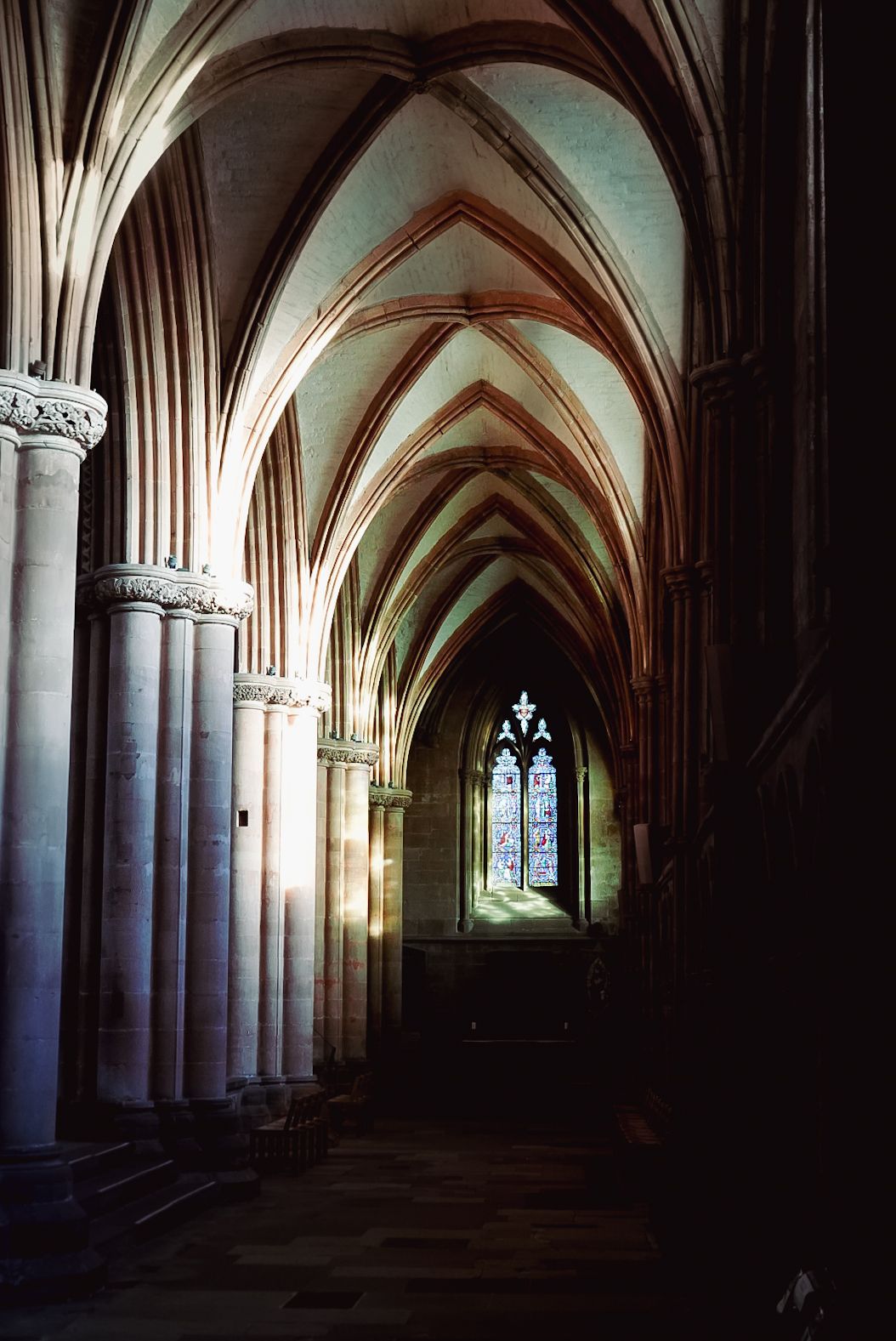 Gothic cathedral nave with ribbed vaulted ceiling, stone columns, and a stained glass window.
