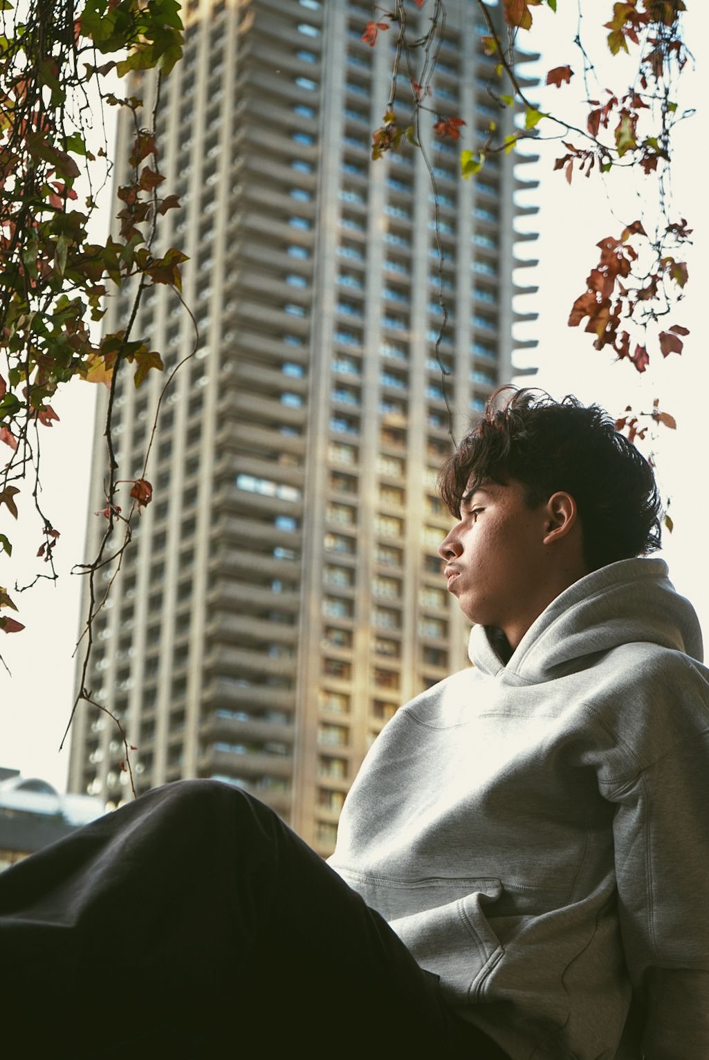 Young man in grey hoodie sits outdoors with tall residential tower block in background.