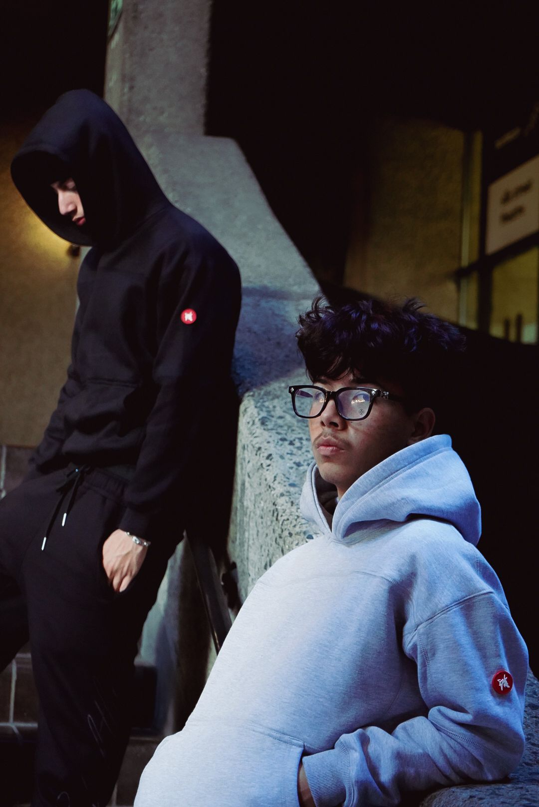 Two young men in black and grey hoodies posing on dark urban staircase at night.