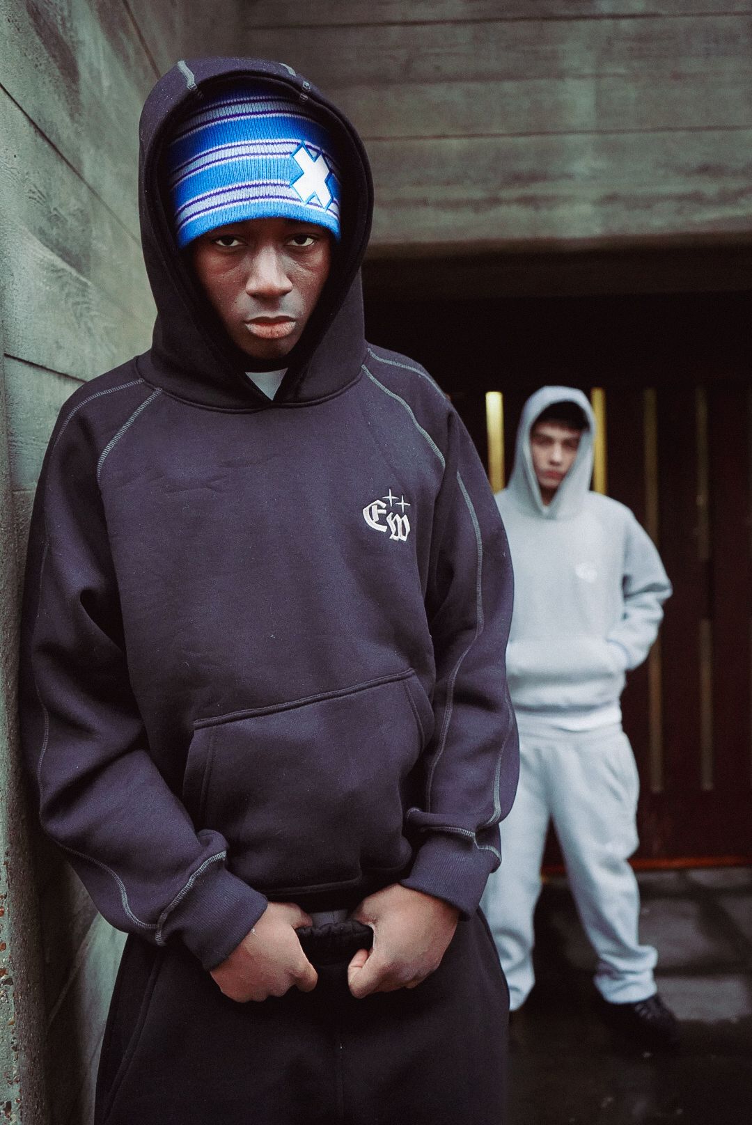 Two young men in hoodies posing in dark urban alleyway, one wearing blue striped beanie hat.