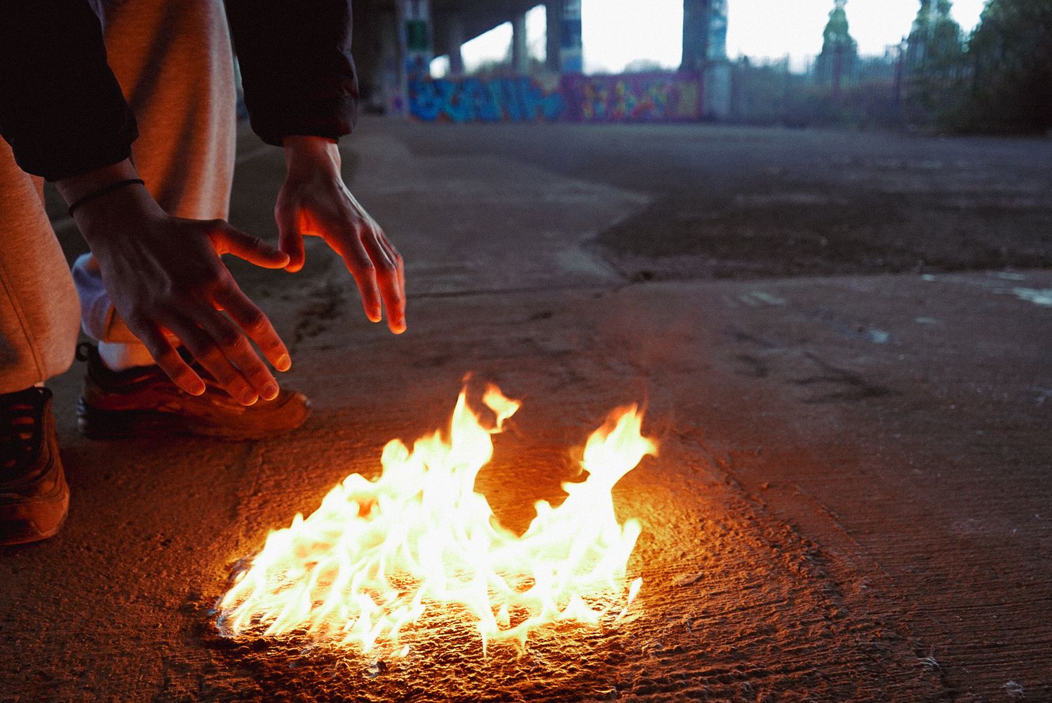 Hands warming over an open fire burning on dark asphalt under a graffiti-covered bridge.