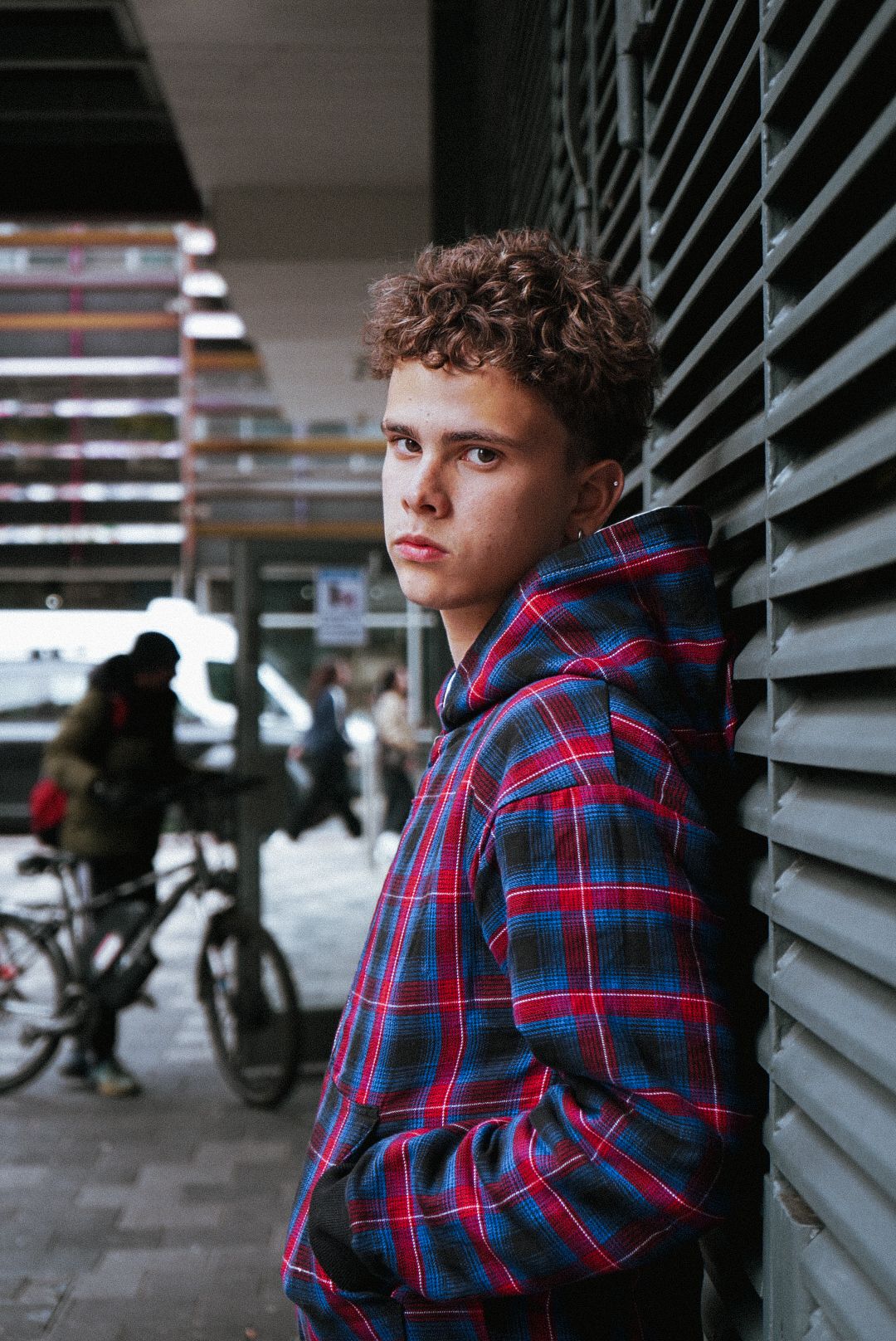 Young man in red and blue plaid hoodie leans against metal shutters in an urban street setting.