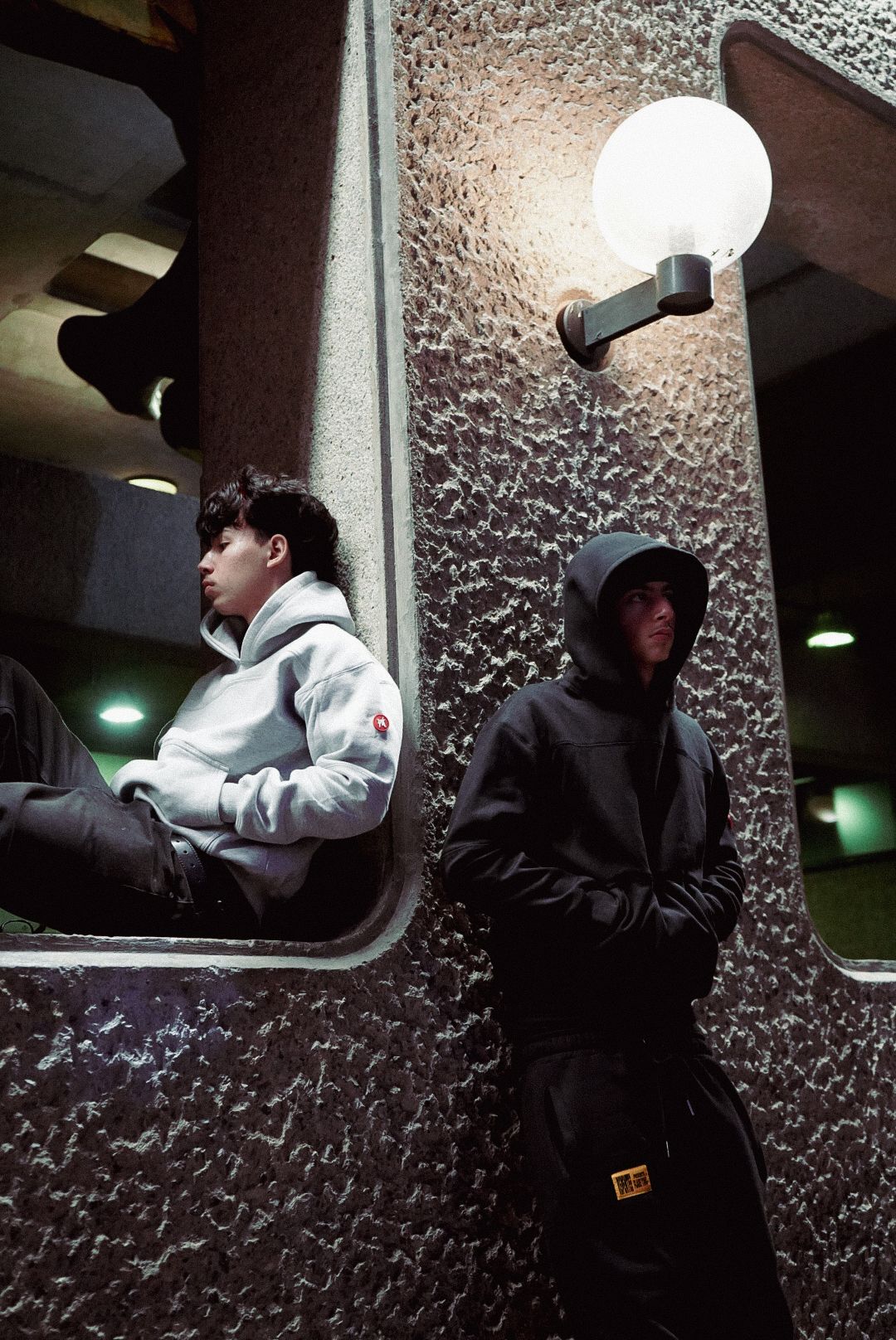 Two men in hoodies stand near a textured wall under a globe light in a dark urban setting.