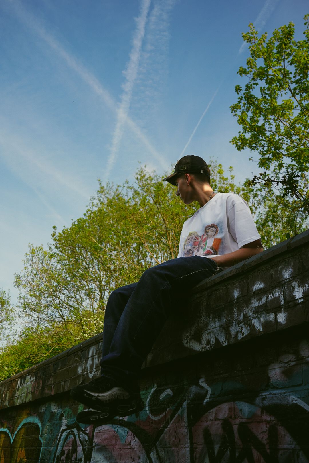Young man in white graphic tee and black cap sitting on graffiti wall under blue sky.