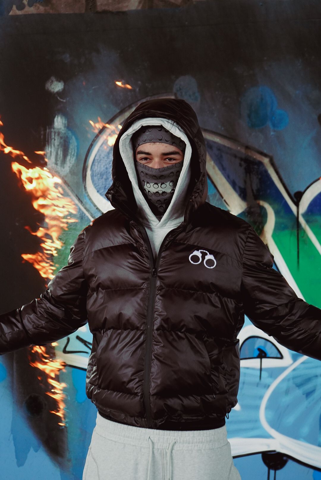 Person in black puffer jacket and face mask posing with fire effect in front of graffiti wall.