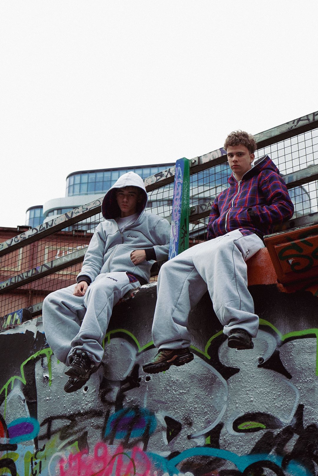 Two young men in hoodies and baggy pants sit on a graffiti-covered wall in an urban setting.