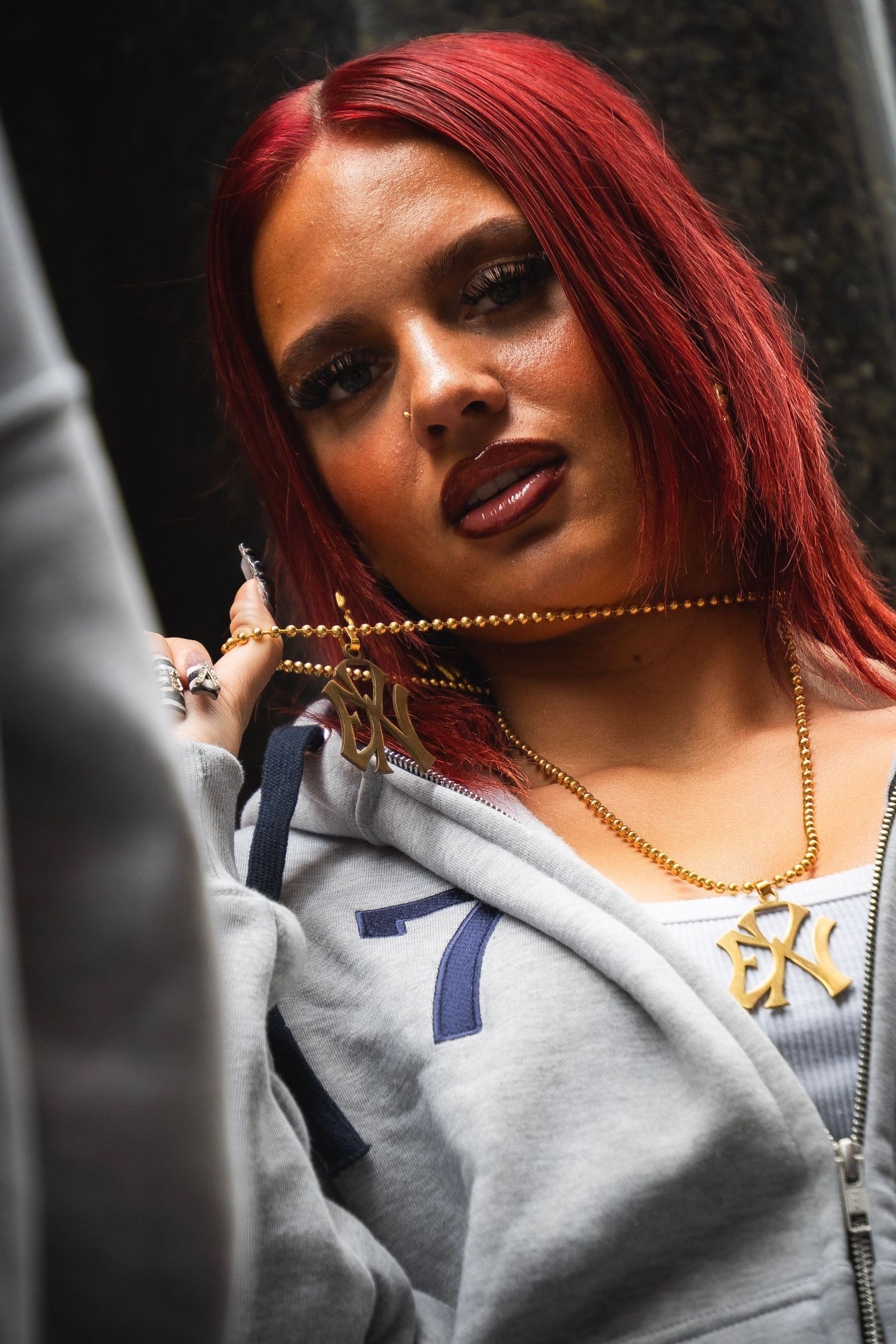 Young woman with red hair wearing gold chain necklaces and a gray hoodie, posing confidently outdoors.