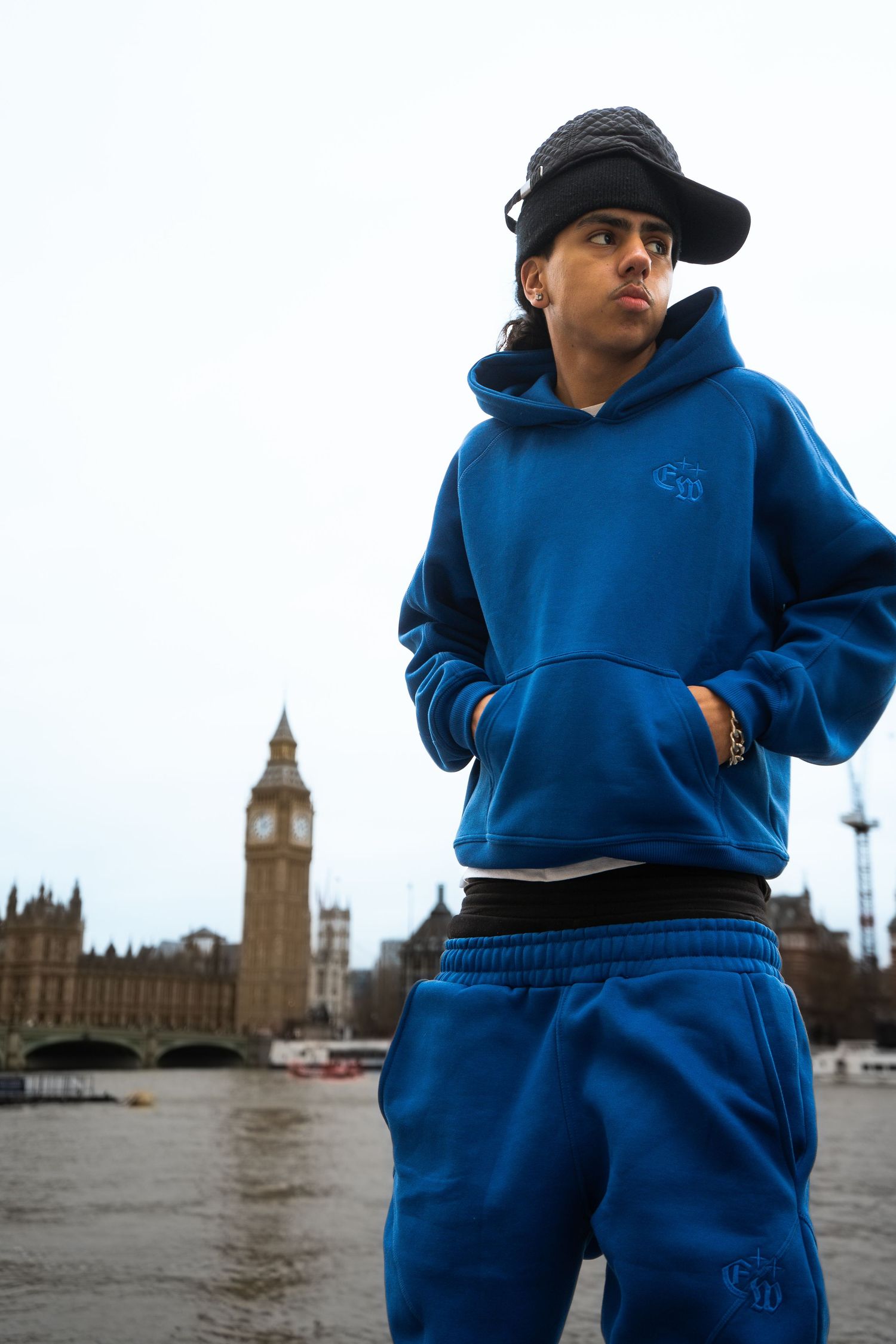 Young man in blue hoodie and sweatpants poses in front of Big Ben landmark in London.