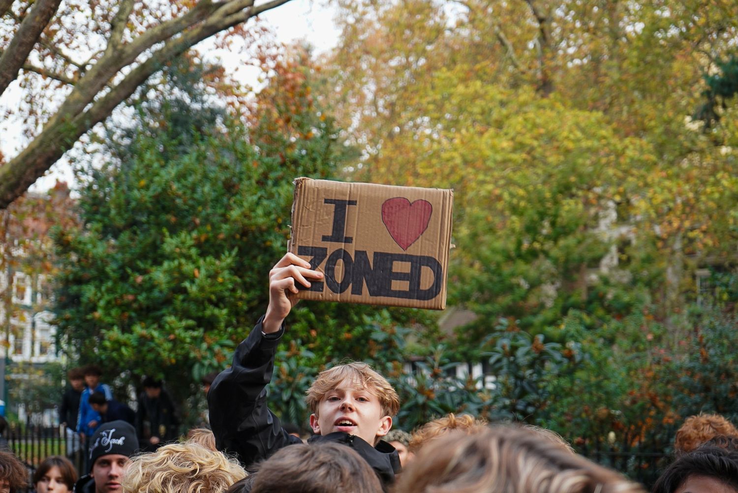 A crowd holds a cardboard sign reading I Love Zoned at an outdoor gathering surrounded by autumn trees.