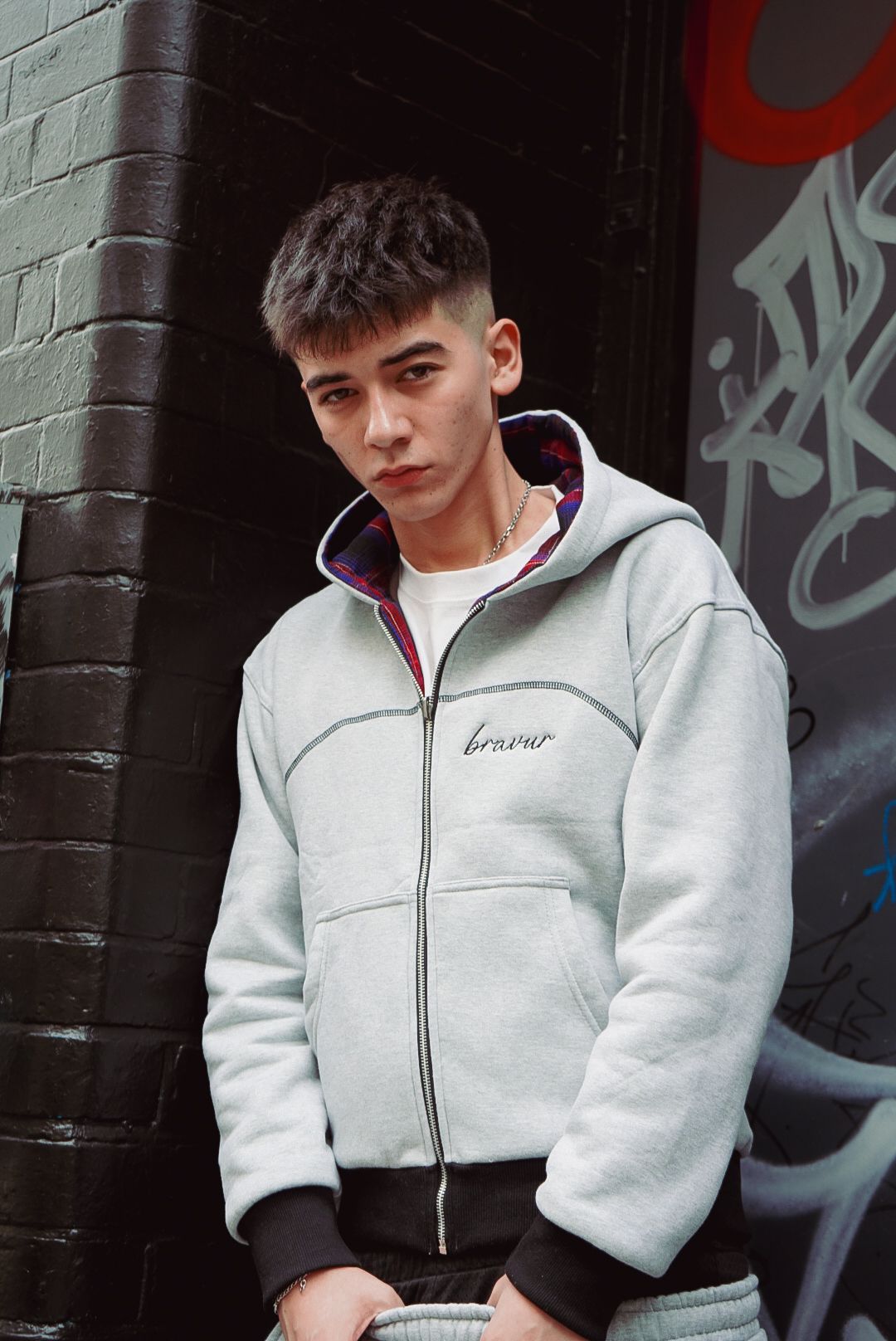 Young man wearing a grey Bravur zip-up hoodie poses against a dark brick wall with graffiti.