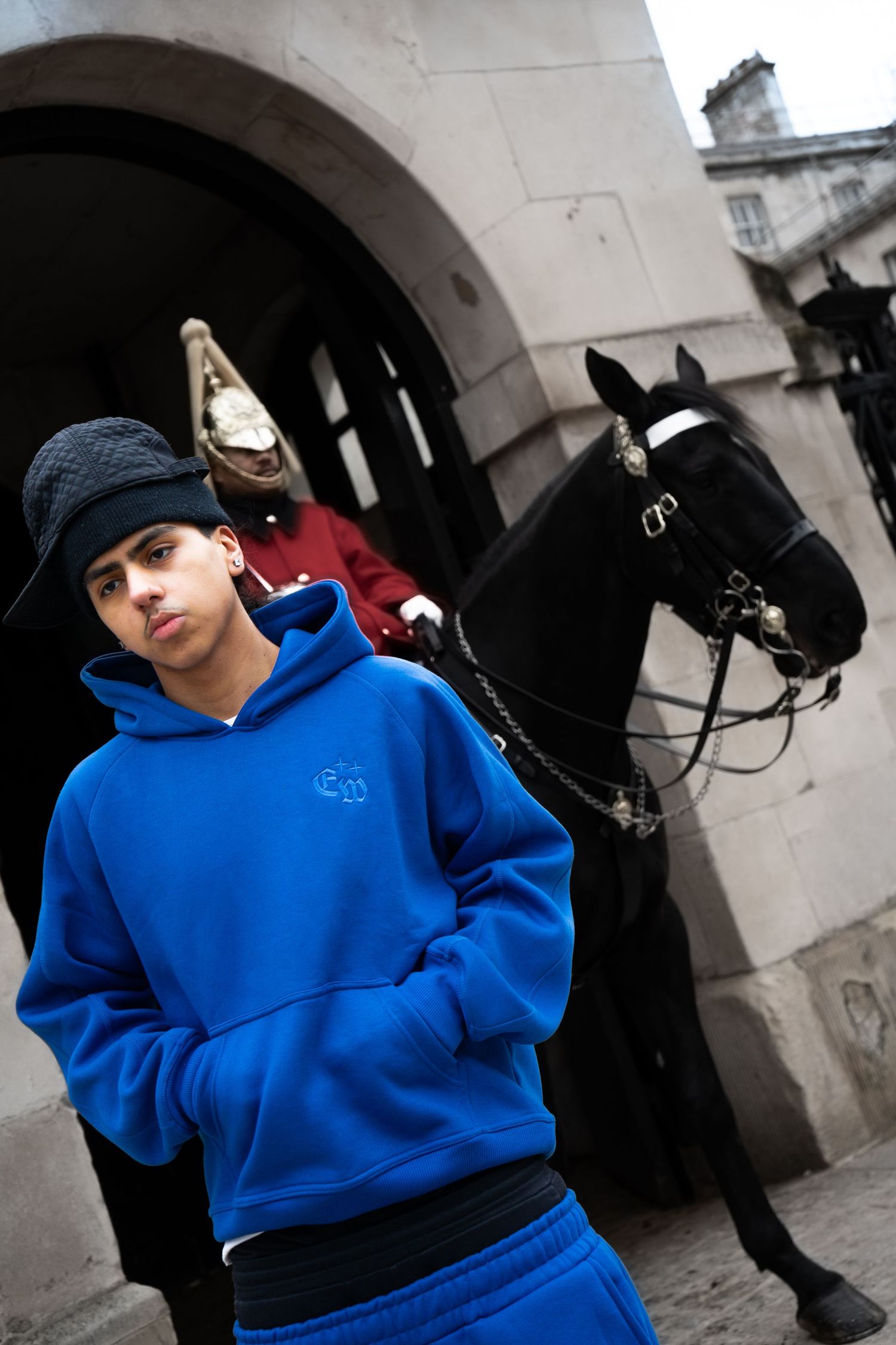 Young man in blue hoodie and beanie standing before Horse Guards with soldier and horse.