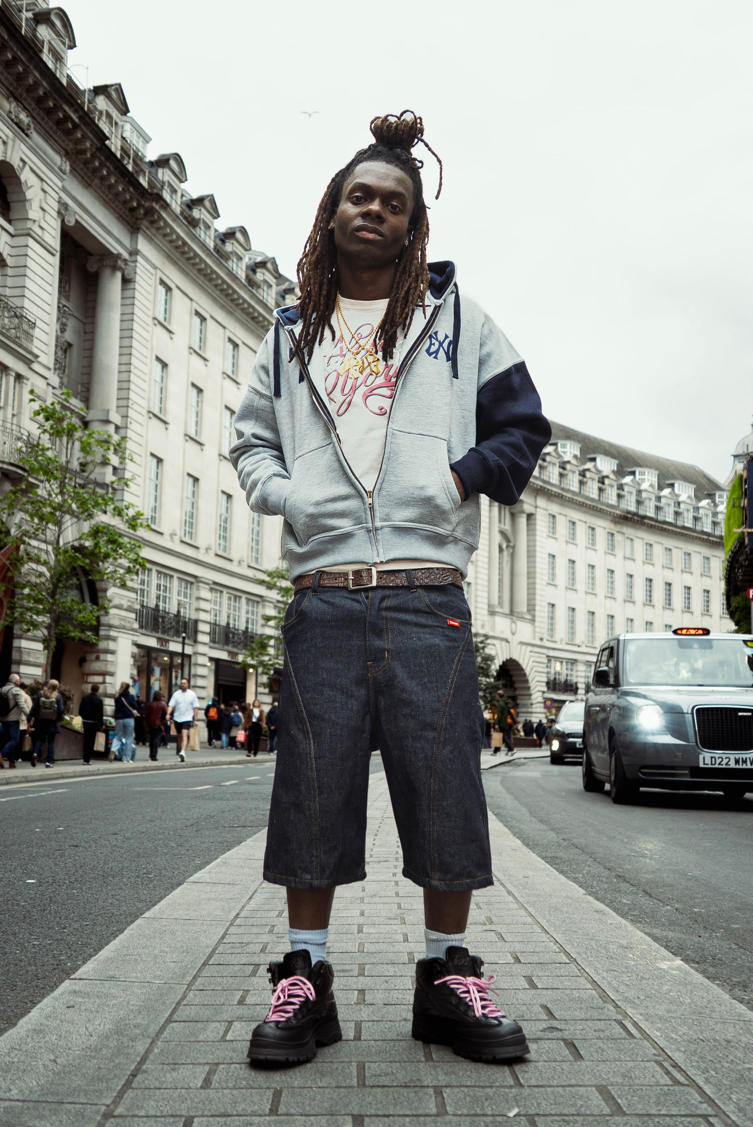 Young man with dreadlocks wearing a grey hoodie, denim shorts, and platform sneakers standing on a London street.