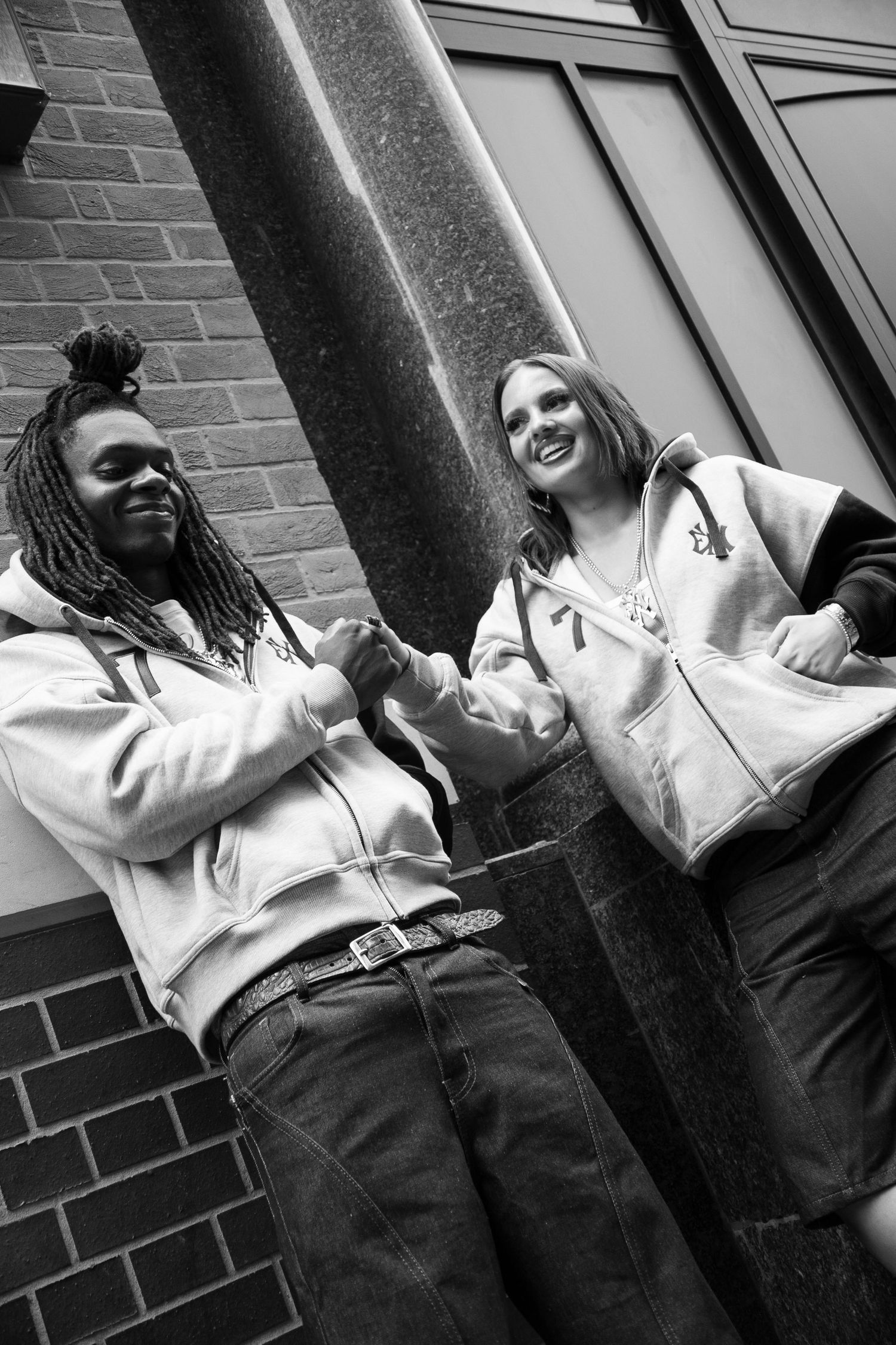 Black and white photo of two smiling friends in hoodies fist bumping outside a brick building with columns.