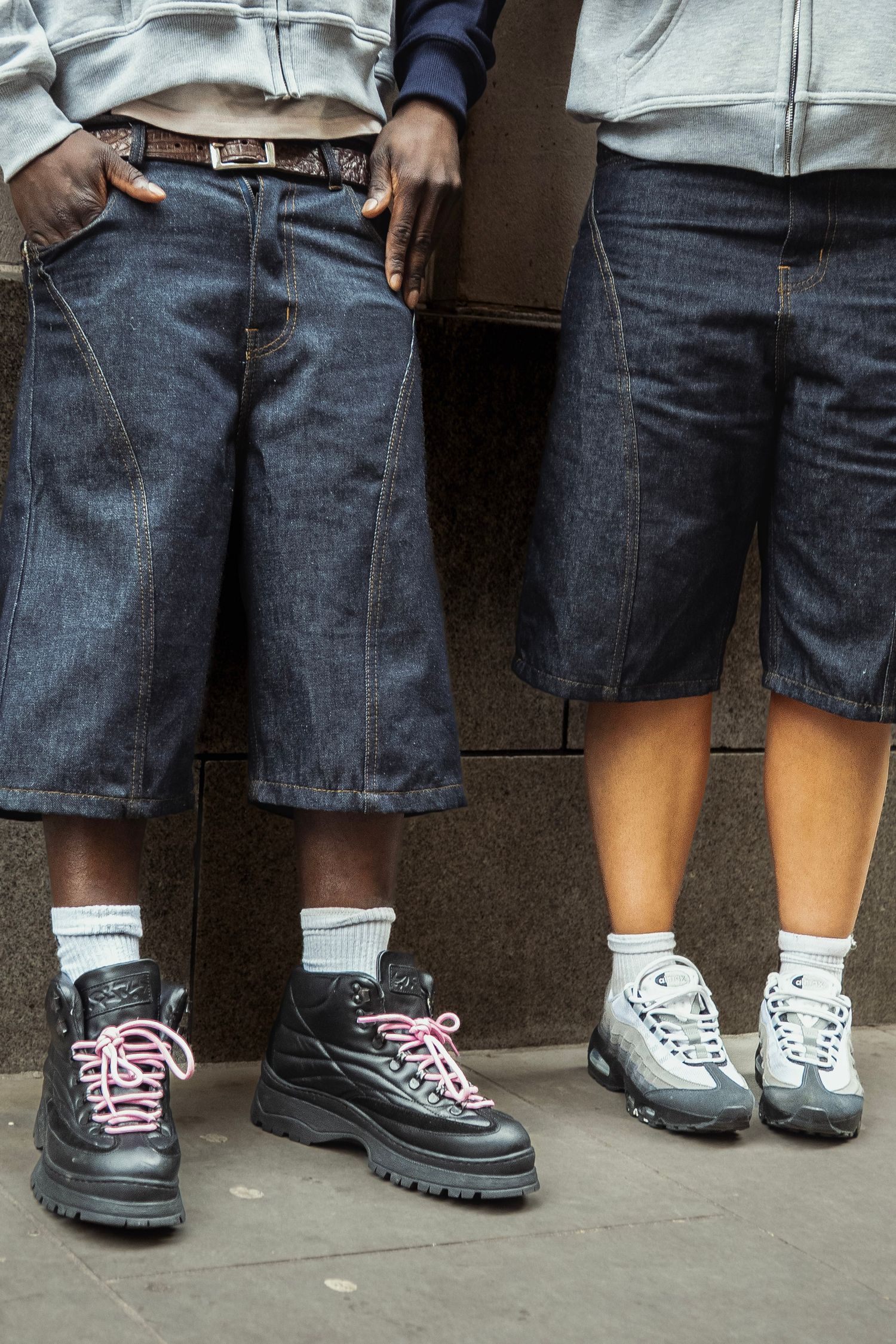 Two people wearing baggy denim shorts, white socks, and chunky sneakers standing against a wall.