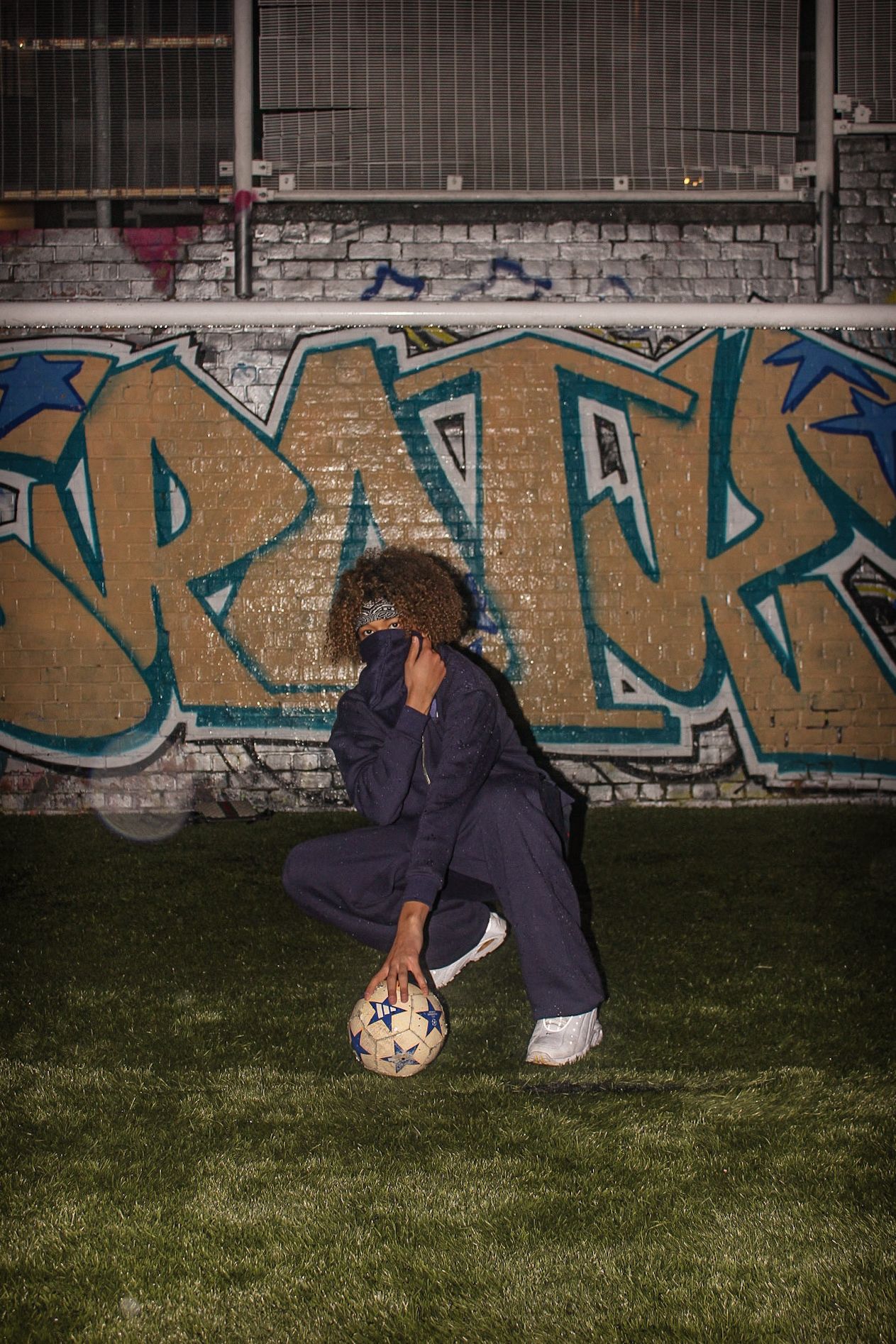 A person in a navy tracksuit crouches with a soccer ball in front of a graffiti-covered brick wall.