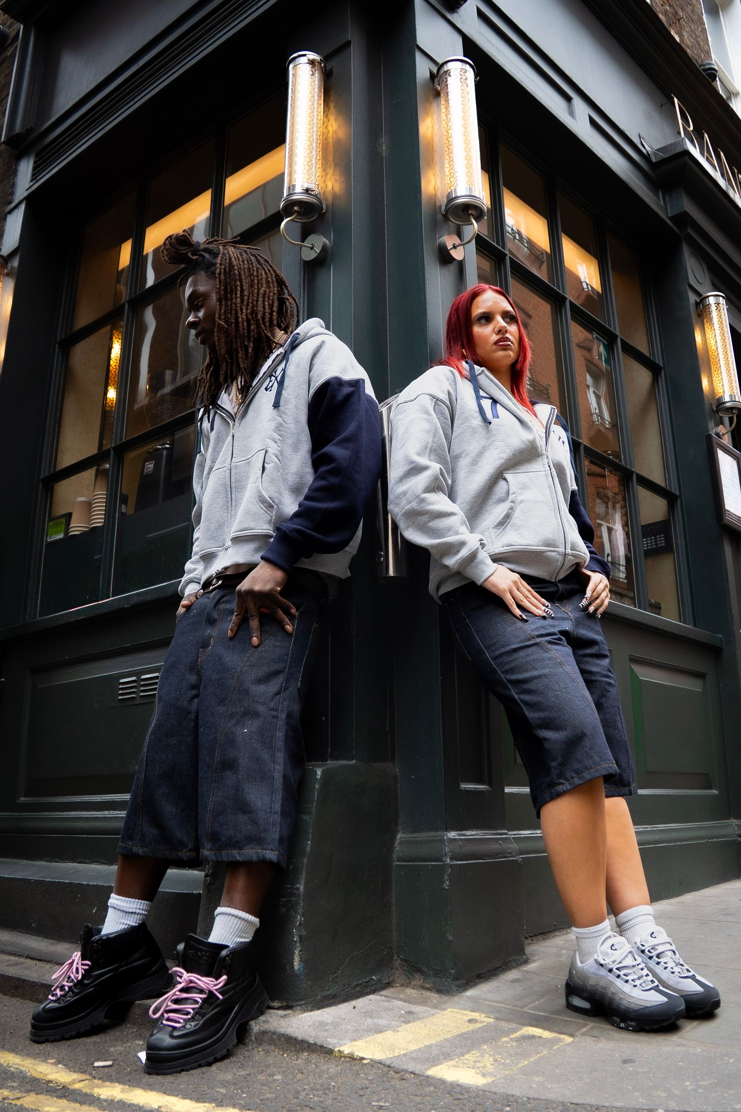 Two models in matching grey zip-up hoodies and denim shorts pose back-to-back outside a dark green storefront.